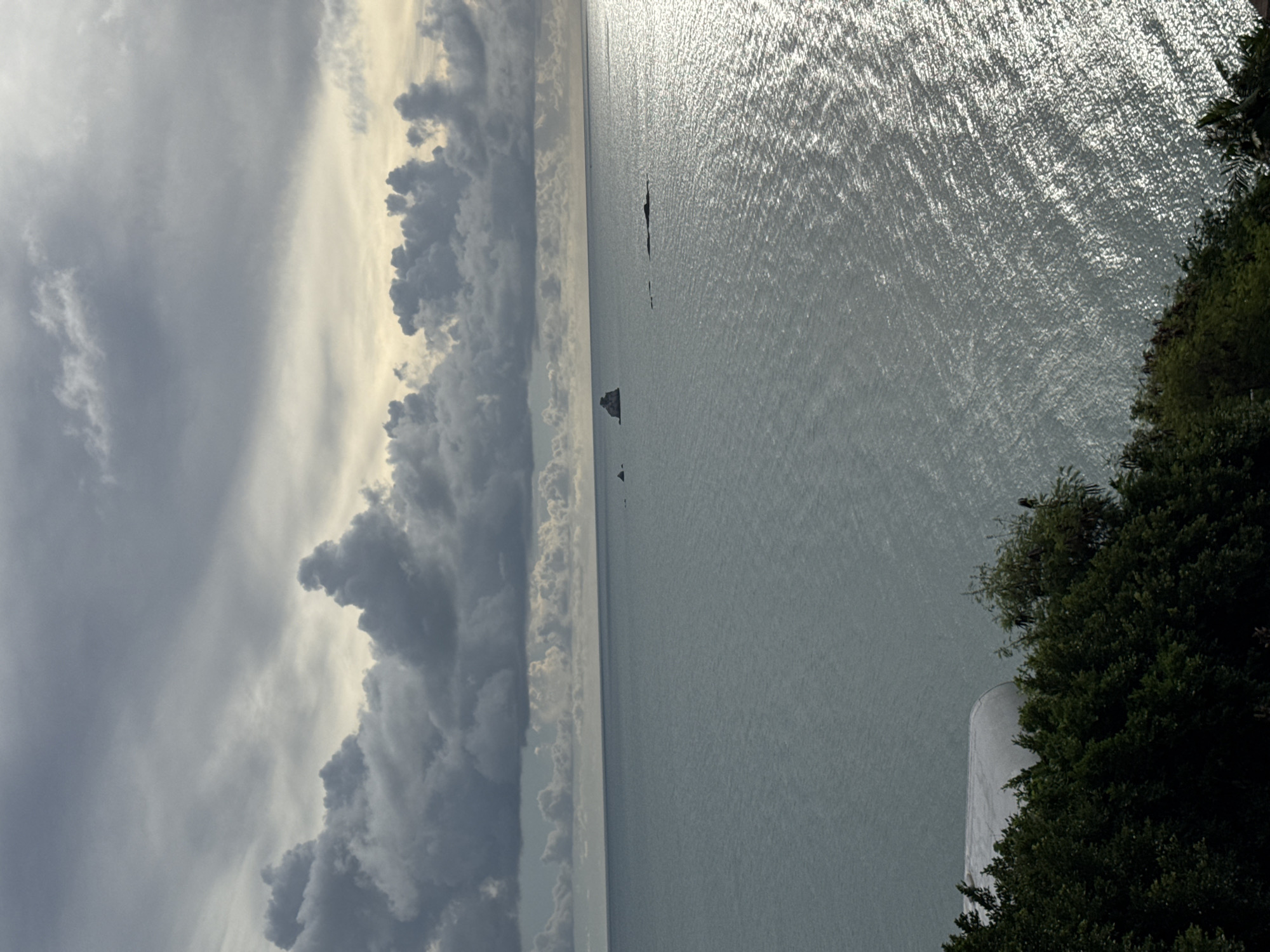The image shows a view of the ocean from a high vantage point, overlooking a coastline covered in dark green foliage. In the foreground, there is a white, curved structure partially visible amongst the trees. The ocean extends to the horizon, with the sun reflecting brightly on its surface, creating a shimmering effect. In the distance, several dark rock formations emerge from the water. The sky is filled with dramatic, dark gray clouds, with patches of lighter sky visible, suggesting a cloudy or overcast day. The scene appears to be a natural landscape, likely a coastal area.