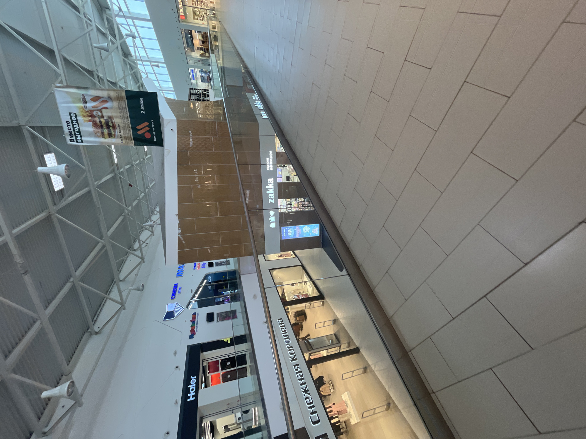 The image displays the brightly lit, multi-level interior of a shopping mall in Ulyanovsk, Russia, captured from a tilted perspective. The scene appears quiet and devoid of people. The mall features a spacious design with a high ceiling supported by a white metal truss structure, integrating skylights that allow ample natural light. The floor is tiled with light-colored rectangular panels.
Storefronts are visible across multiple levels, separated by glass railings. A large banner prominently advertises "ВКУСНО И ТОЧКА" (Vkousno i Tochka), showing a burger and fries, with accompanying text "Вместе выгоднее" (More profitable together), "Биг Хит Комбо" (Big Hit Combo), and "2 этаж" (2nd floor), indicating its location.
Other identifiable store names written in Cyrillic include "Haier," "Снежная Королева" (Snezhnaya Koroleva - Snow Queen), and "zakka," which further states "подарки и аксессуары" (gifts and accessories). In the background, signs for "KUCHEN" and "СИНЕМА" (Cinema) are also discernible, along with "UINVAX". Some store facades are clad in reflective brown or golden panels. The overall impression is a modern, clean, and well-maintained commercial environment.