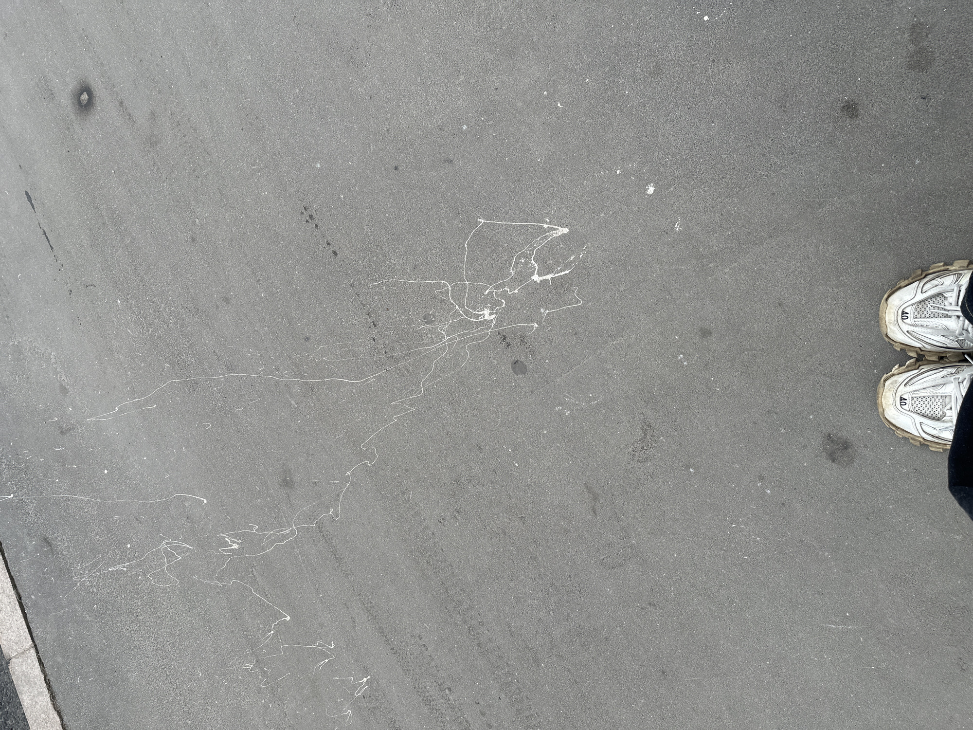 The image shows a section of asphalt pavement, possibly a street or sidewalk in Moscow, Russia, based on common urban elements. Splatters of white paint are irregularly dispersed on the gray surface, extending from the lower left towards the center. In the lower right corner, a pair of white athletic shoes are partially visible, suggesting a person is present but not fully in the frame. Small dark spots and other minor surface imperfections are also visible on the pavement.