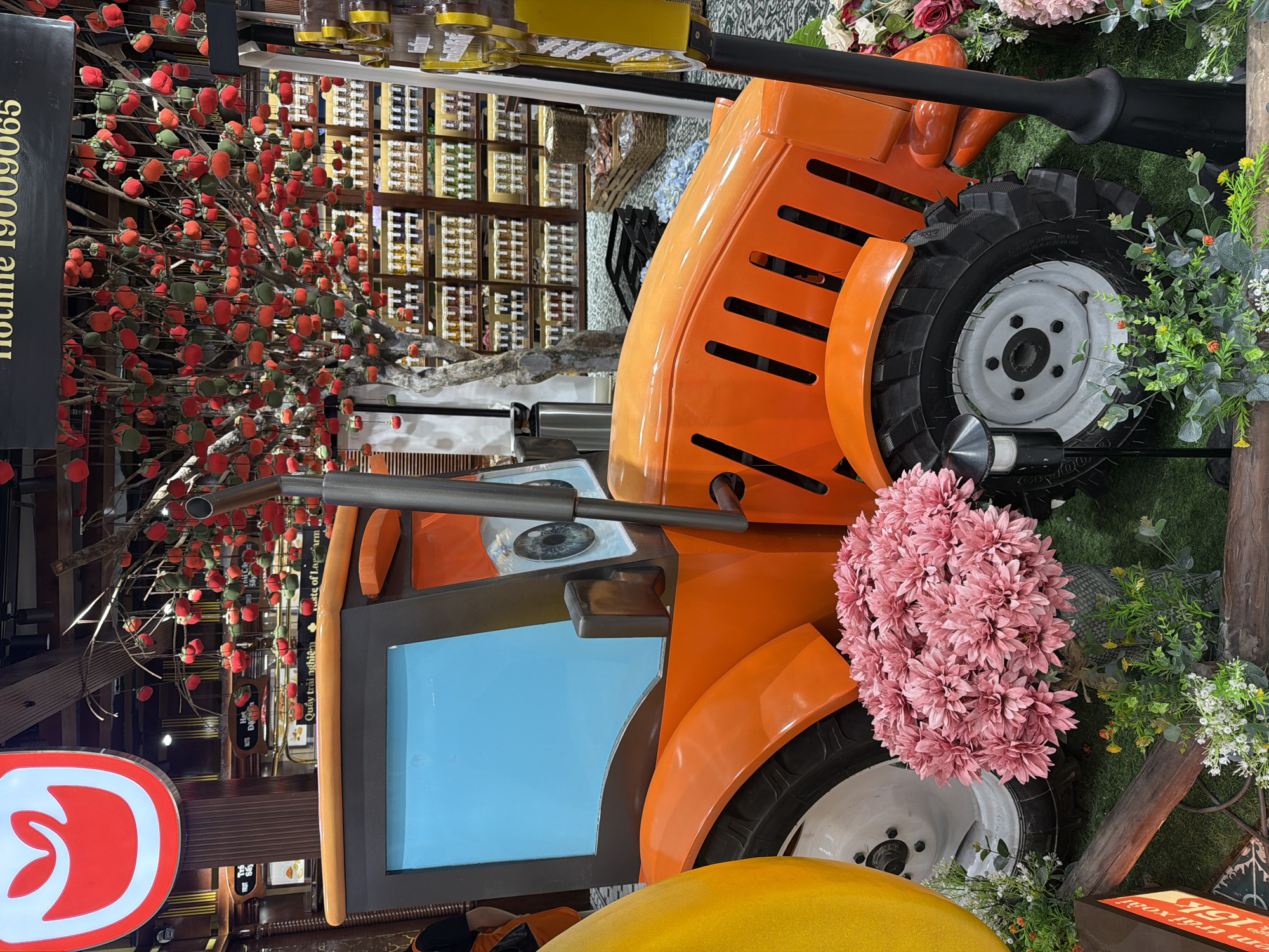 The image captures an outdoor market stall in Da Nang, Vietnam, featuring a vibrant orange toy tractor as a central decorative element. The tractor, with its blue windshield and cartoonish eyes, is adorned with a large bouquet of pink flowers in front of its front wheel. Behind the tractor, a tree branches out, laden with numerous small red and green decorations resembling persimmons. Shelves filled with jars of products are visible in the background, suggesting a retail setting. The ground is covered in artificial green turf, enhancing the market's playful aesthetic. The scene appears to be a promotional display, possibly for agricultural or food-related products, given the tractor and fruit-like decorations.