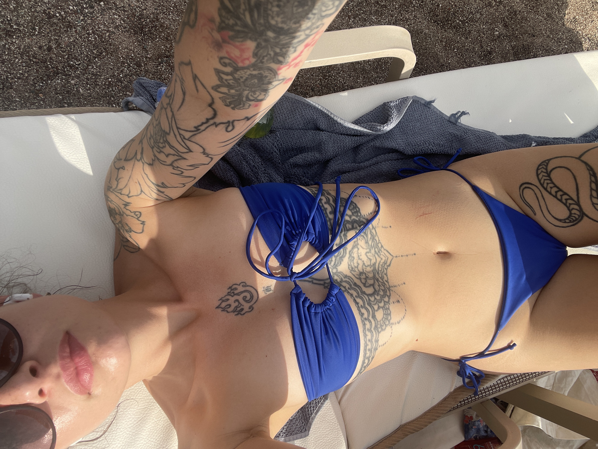 An overhead self-portrait captures an individual, appearing female, reclining supine on a white padded sun lounger, positioned on a pebble beach in Rafailovići, Montenegro. The person wears a royal blue two-piece bikini; the top is a strapless bandeau style with central gathering and ties, and the bottom is a low-rise string-tie brief. Dark sunglasses cover the upper half of the face, and a white wireless earbud is visible in the right ear.
Numerous tattoos are present: a stylized flame symbol with a small character below it on the sternum, an intricate mandala-style pattern across the upper abdomen, complex floral and organic designs on both arms (with a distinct rooster-like figure on the right upper arm), and a coiled snake on the right thigh.
Strong sunlight illuminates the scene, casting defined shadows. A dark blue or grey towel is draped over the sun lounger to the right of the individual. The ground consists of dark, small pebbles or coarse gravel. A partial view of a second sun lounger is visible on the left, and a small, dark plastic bottle is partially obscured on the ground near the right lounger. The overall scene depicts a person relaxing on a sunny beach.