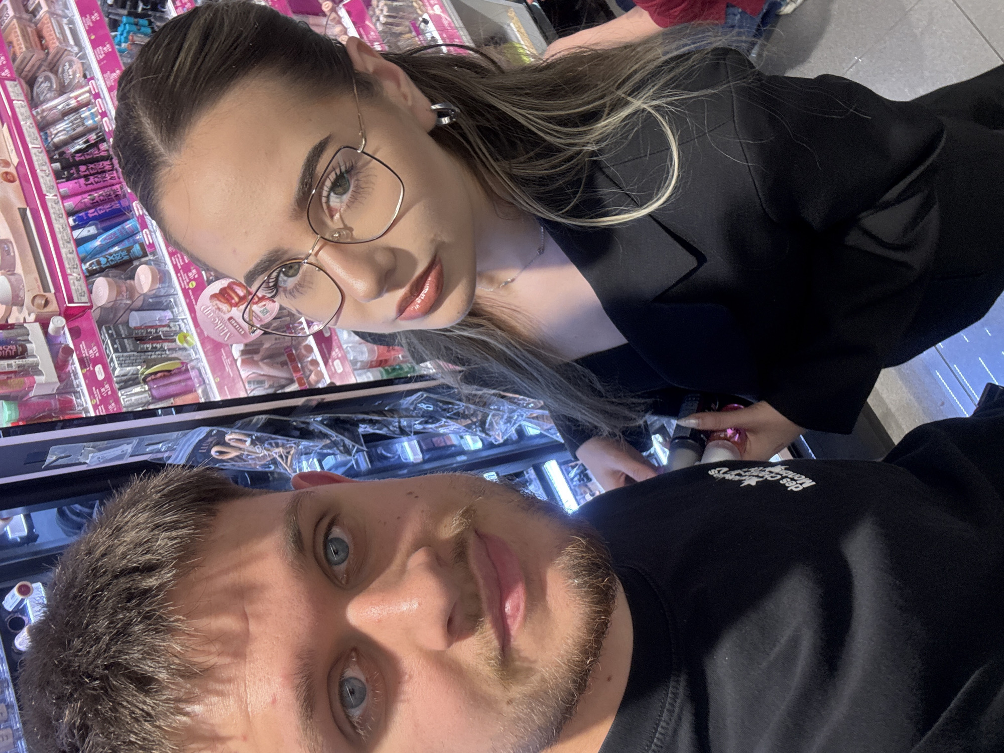 A young man and woman are pictured in an apparent selfie taken at a store. The woman, wearing glasses and a black blazer, is looking down at the camera with a slight smile, while the man, wearing a black t-shirt, is positioned below her. The store appears to sell cosmetics, with shelves of various products visible in the background. The location appears to be in Blagoevgrad, Bulgaria.