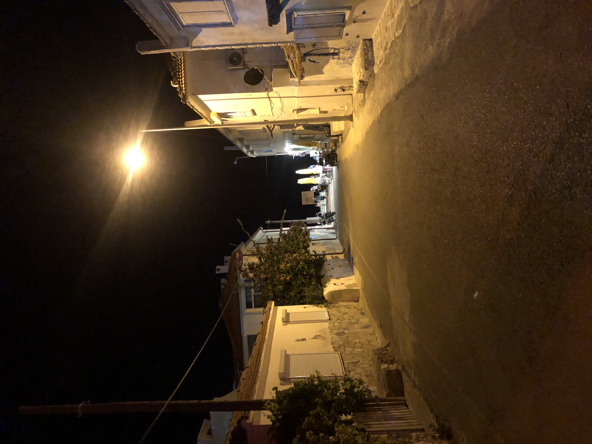 An eye-level, medium shot captures a street scene at night in Karaburun, Turkey, illuminated by a streetlamp. The camera is positioned in the middle of a paved street that recedes into the darkness. Buildings line both sides of the street. On the left, a building with a stone base and white upper walls is visible, partially obscured by foliage. To the right, a row of white buildings with tiled roofs stretches along the street. In the distance, a few figures can be seen, along with parked motorcycles and tables with umbrellas, suggesting an outdoor seating area. The sky is dark, with the bright glare of the streetlamp dominating the upper right portion of the frame. The overall impression is of a quiet, dimly lit street in a coastal town at night.