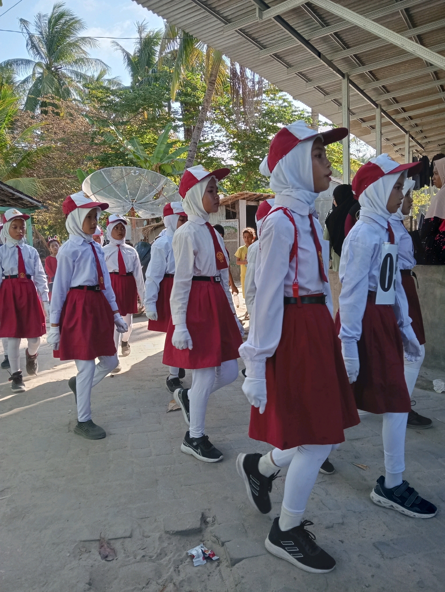 In Pagerungan Kecil, Indonesia, a group of young girls in uniform are participating in a parade. The girls are wearing white shirts, red skirts, white hijabs, red and white hats, and white gloves. Some have red ties and suspenders. They are walking on a dirt road lined with trees and a building with a corrugated metal roof. A satellite dish is visible in the background. The scene suggests a school event or a community celebration.