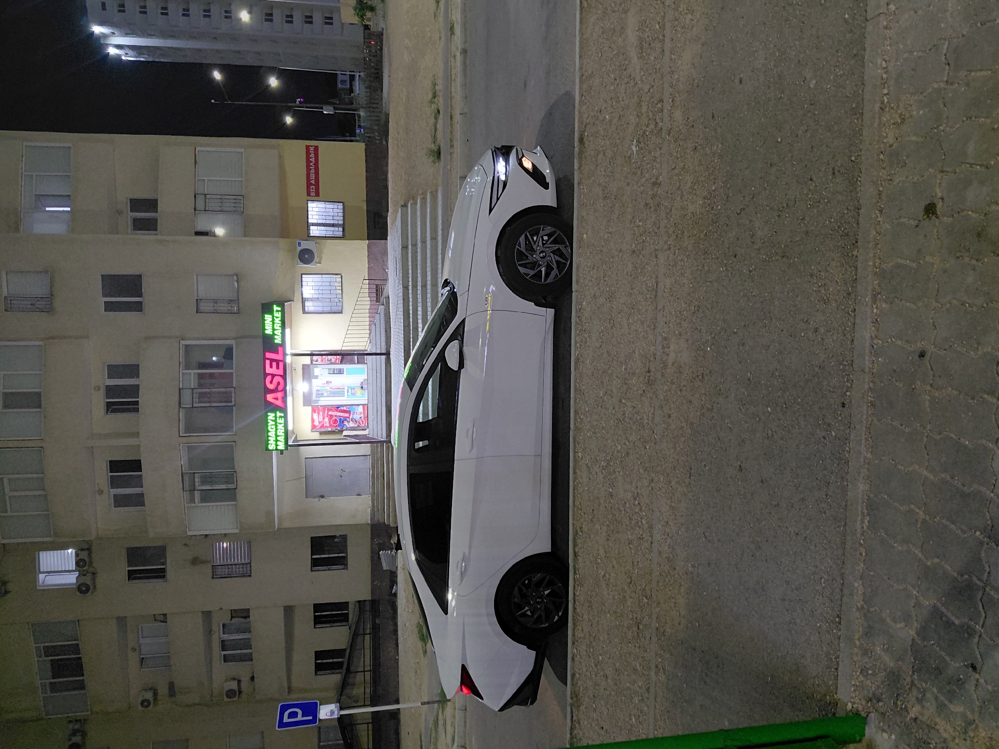 The image shows a white sedan parked on the side of a street in front of a multi-story building at night. The building has a storefront with neon signs that read "SHAGYN MARKET" and "ASEL MINI MARKET". The car is a modern sedan with its headlights on. The street appears to be paved with asphalt, and there is a sidewalk made of interlocked concrete pavers. The building is illuminated, and some windows are lit, suggesting occupancy. The overall scene depicts a typical urban street in Aktau, Kazakhstan, at night.