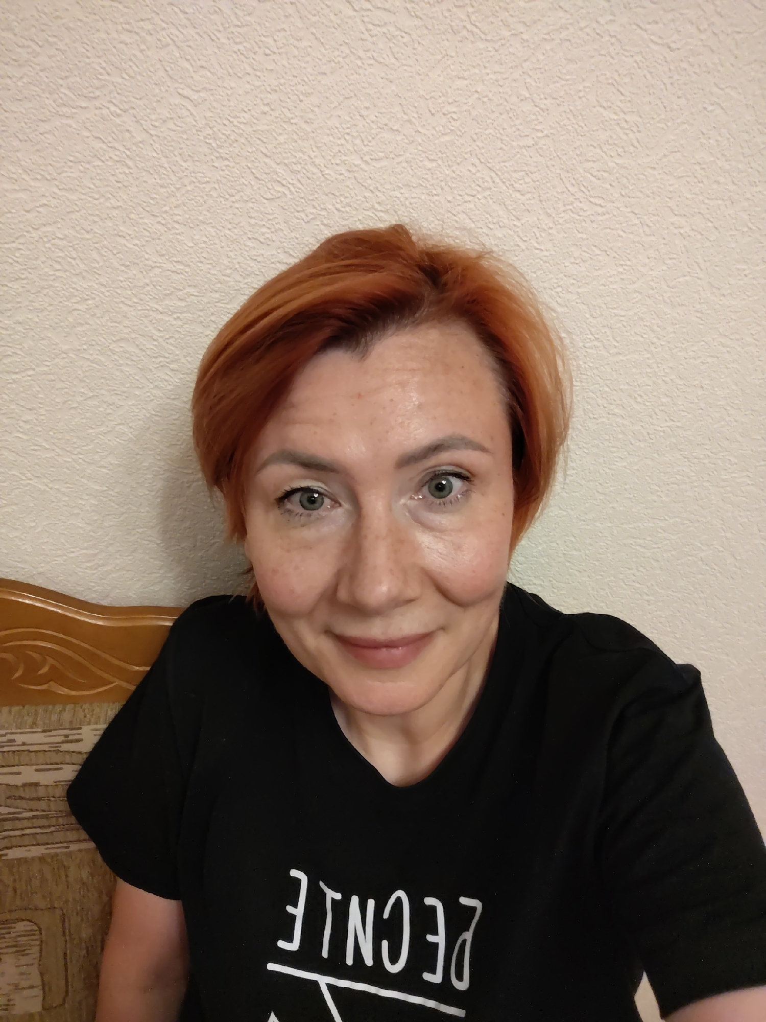 The image shows a close-up, selfie-style portrait of a woman with short, reddish-orange hair and fair skin with freckles. She is wearing a black t-shirt with white lettering on it, some of which is visible and appears to be in reverse. She has light-colored eyes and is smiling slightly. The background is a textured, light-colored wall. The woman is positioned in what appears to be an indoor setting, possibly a home, with a portion of a wooden headboard of a bed or sofa visible on the left side of the frame. There is no discernible location within the city of Konakovo, Russia, that can be identified from the image.