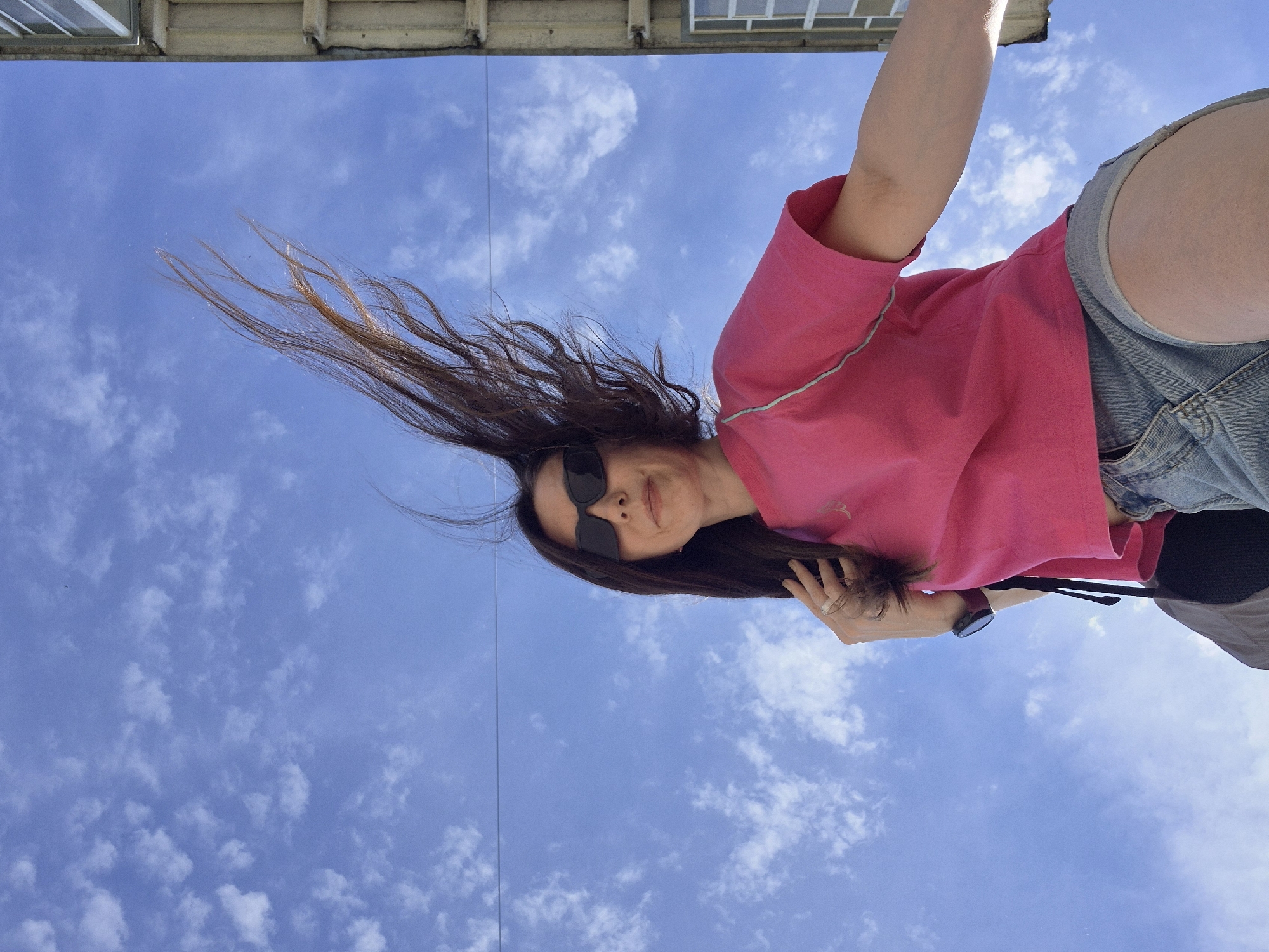A woman wearing sunglasses, a pink t-shirt, and denim shorts is positioned at the bottom-right of the frame, with her head and hair extending diagonally toward the top-left. Her hair is long and brown, blowing in the wind. She is wearing a wristwatch on her left wrist and several rings on her fingers. She appears to be looking into the camera. The background is a bright blue sky filled with scattered white clouds. A building overhangs the top of the frame. The location is outdoors.