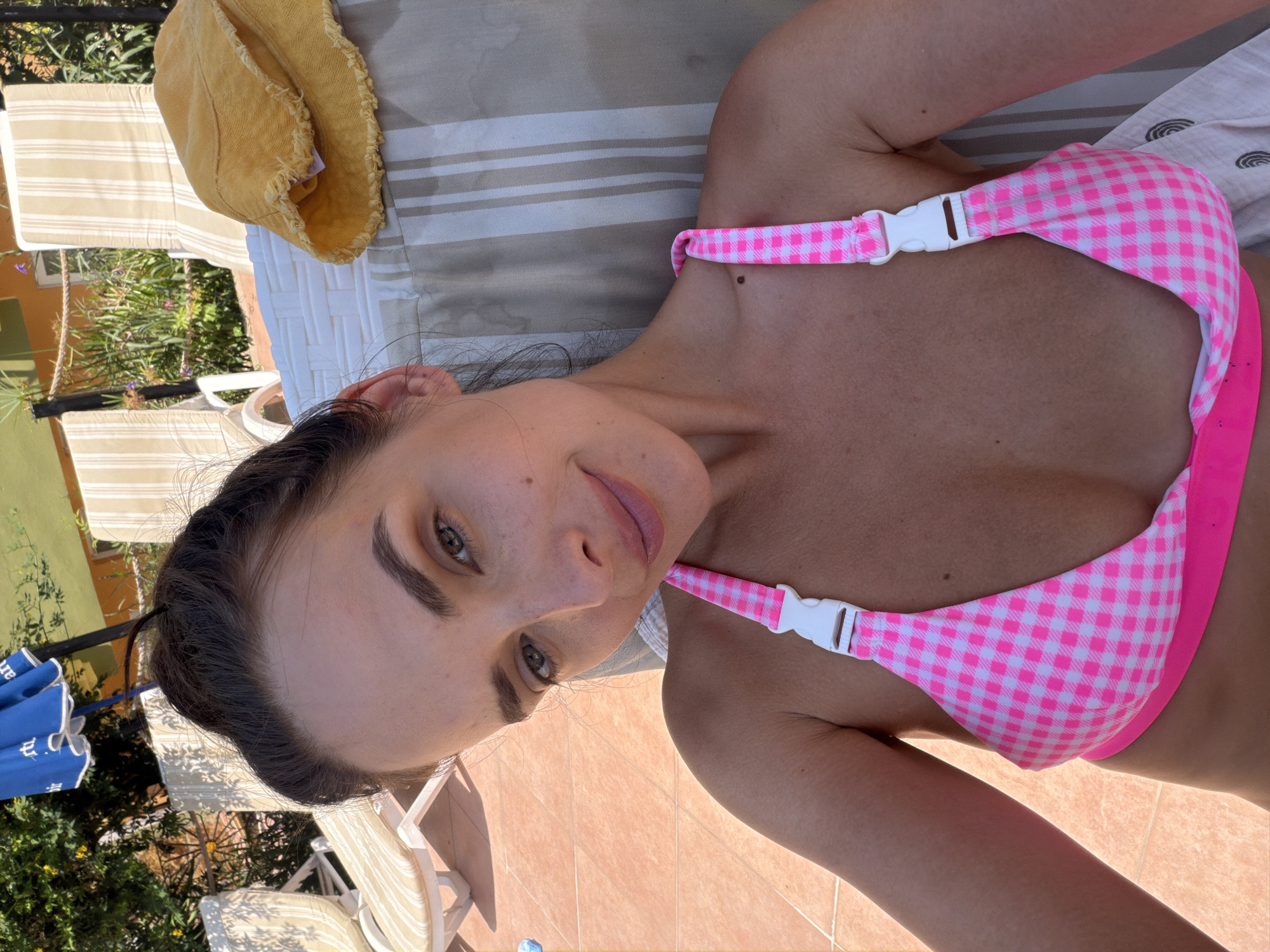 A woman with dark hair pulled up is lying on a lounge chair outdoors, wearing a pink and white checkered bikini top. She is looking towards the camera with a slight smile. Behind her, lounge chairs and a yellow hat are visible. The ground appears to be tiled and reddish-orange. Sunlight illuminates the scene. The location is stated to be in Fethiye, Turkey.