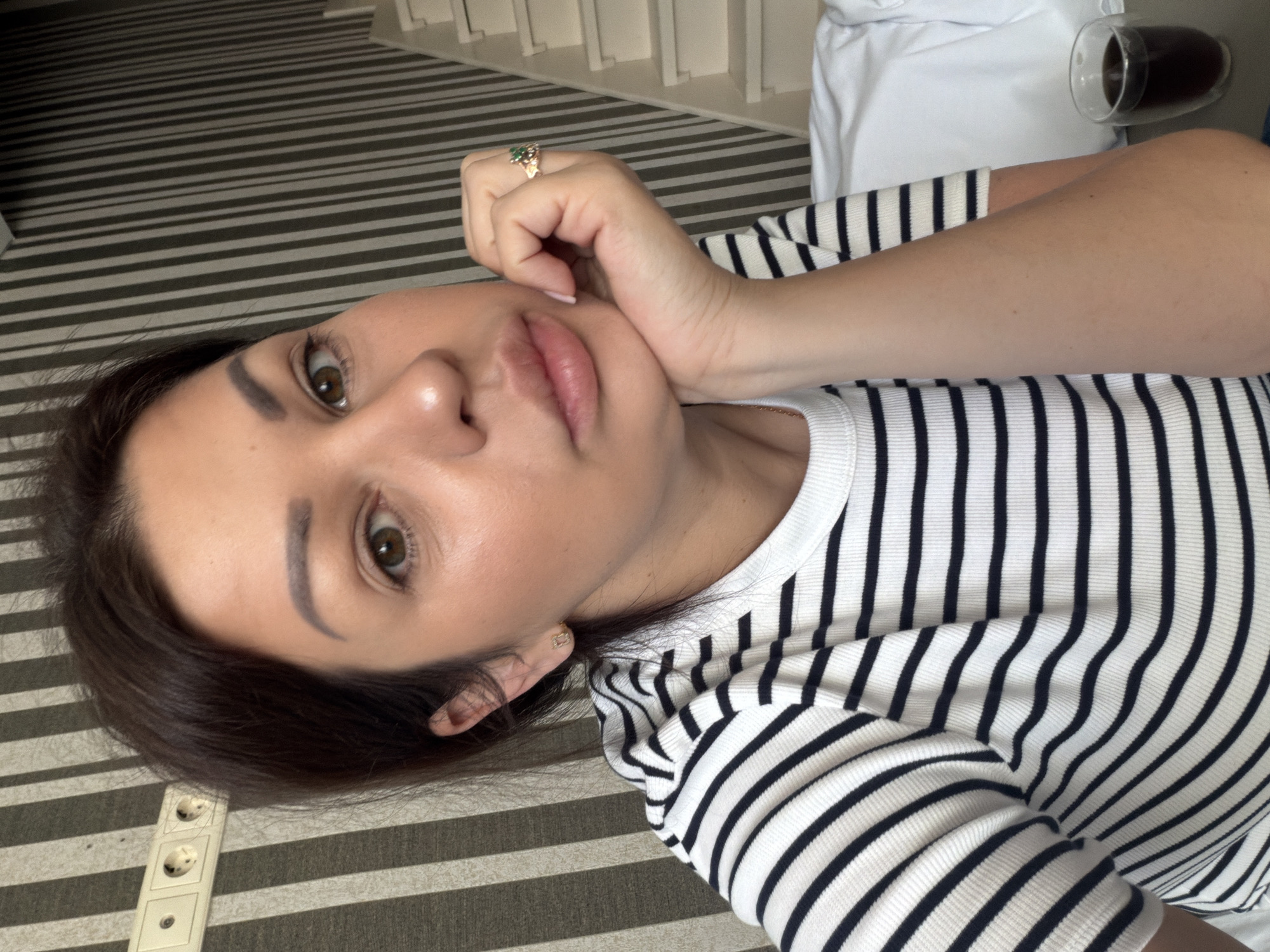 The image shows a close-up, waist-up shot of a woman with dark hair and light brown eyes, wearing a black and white striped shirt. She is resting her chin on her hand, which is adorned with a ring on her right index finger. Her left hand is visible near her neck. The background consists of a striped wallpaper and a white staircase, suggesting an indoor residential setting. A glass of dark liquid, likely coffee, is visible on a surface to her right. The presence of a power outlet on the wall to her left further indicates an indoor, possibly residential or hotel, environment. The overall lighting suggests the photo was taken during the daytime. The context of Sochi, Russia, would imply this setting is within a dwelling or accommodation in that city.