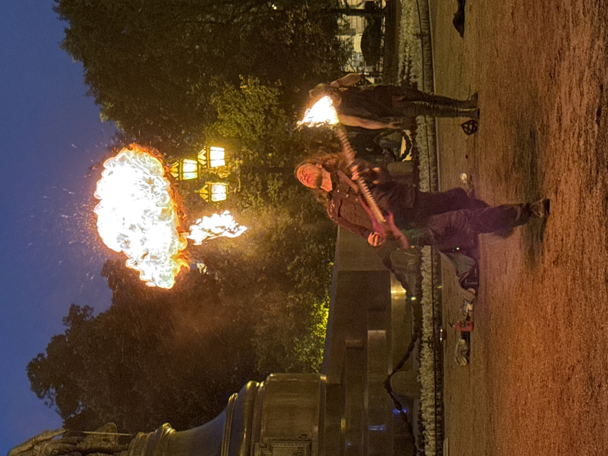 The image depicts a fire performance taking place at night in a public square, likely in Saint Petersburg, Russia, given the architectural style and presence of historical monuments and street lamps. In the foreground, a man with long, dark hair is playing a guitar while breathing fire, creating a large, illuminated plume of flames above his head. He is wearing dark clothing and is in motion, leaning back with his mouth open, exhaling the fire. To his right and slightly behind him, another person is visible, also with fire around their head, though their actions are less clear.
The background features a large, ornate monument on the left, possibly a statue on a pedestal, illuminated by external lighting. Behind the performers, several traditional street lamps with multiple glowing bulbs cast a warm light. Trees with dense foliage surround the performance area, and the ground is paved with a reddish-brown material, likely gravel or dirt. The sky is dark blue, indicating nighttime. Scattered on the ground are various objects, including what appears to be a fire extinguisher, bags, and other performance equipment. The scene suggests an outdoor evening event, possibly a festival or street entertainment.