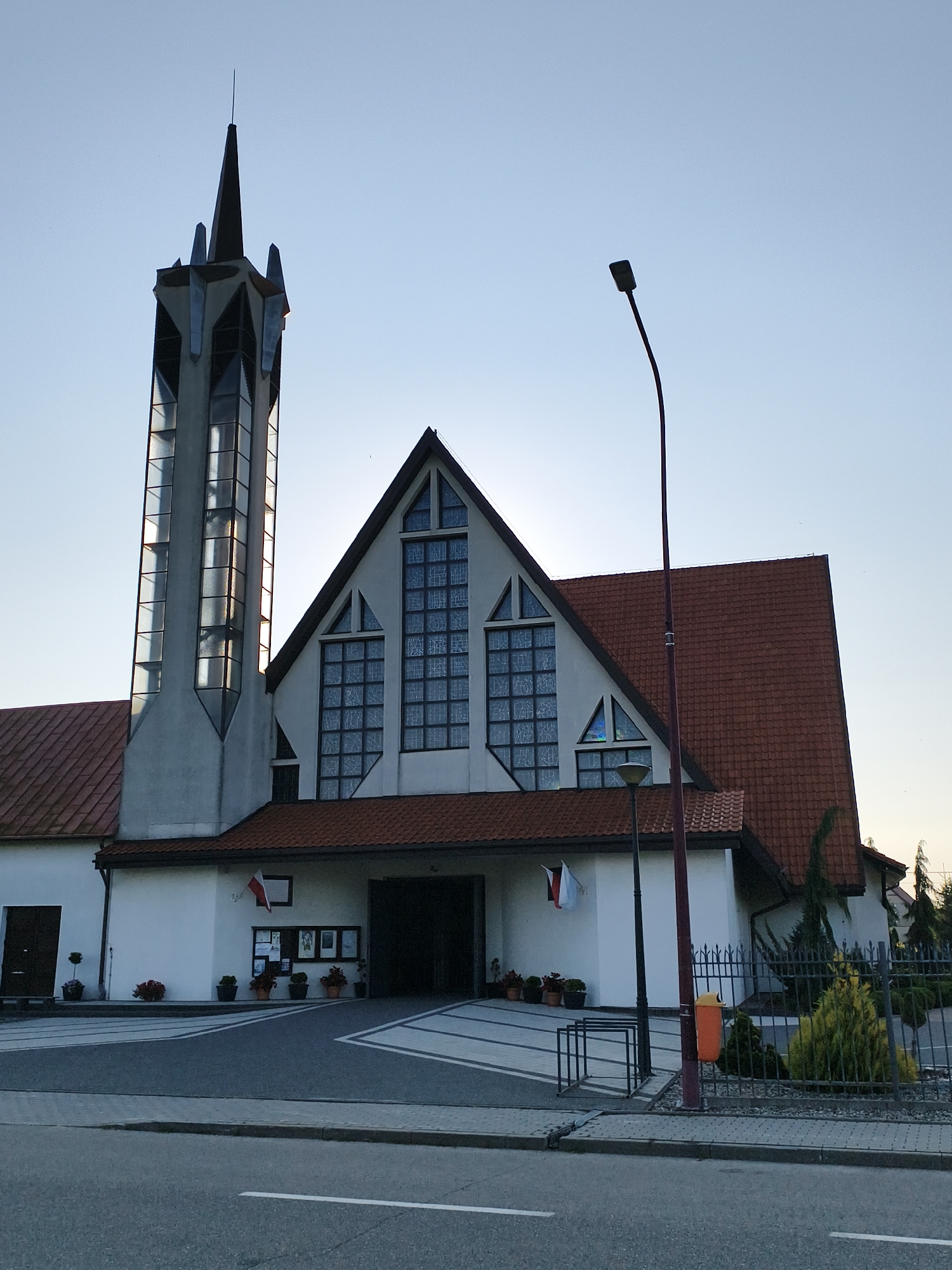 The image shows a modern church building with a tall, angular tower featuring a pointed spire. The main body of the church has a steeply pitched, red-tiled roof and large, stylized stained-glass windows. A wide, paved entrance area is visible, flanked by potted plants and notice boards. A Polish flag is displayed near the entrance. A street runs in front of the church, with a sidewalk and a lamppost. The sky is clear and blue, suggesting it is either early morning or late afternoon. This is the Church of Our Lady of Sorrows in Tczew, Poland.