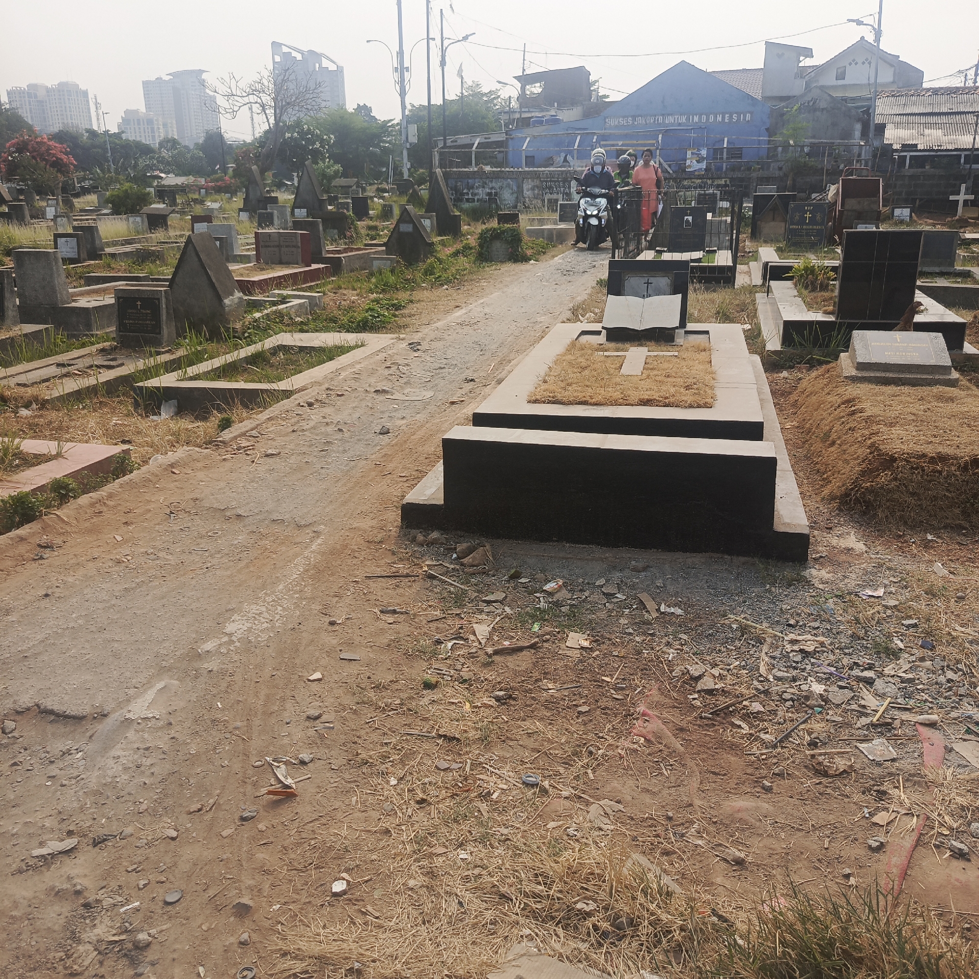 The image depicts a cemetery in South Jakarta, Indonesia, characterized by numerous graves and a sandy, unpaved path. In the foreground, a prominent dark stone tomb with a stylized cross design is visible. Beyond this, a motorcycle carrying two individuals, one a person in an orange shirt, is moving along the path. The background features a blue building with text that appears to be a slogan or name, likely related to a local organization or business, and residential buildings and high-rise structures further in the distance. The cemetery grounds are dry and somewhat overgrown with grass and debris.