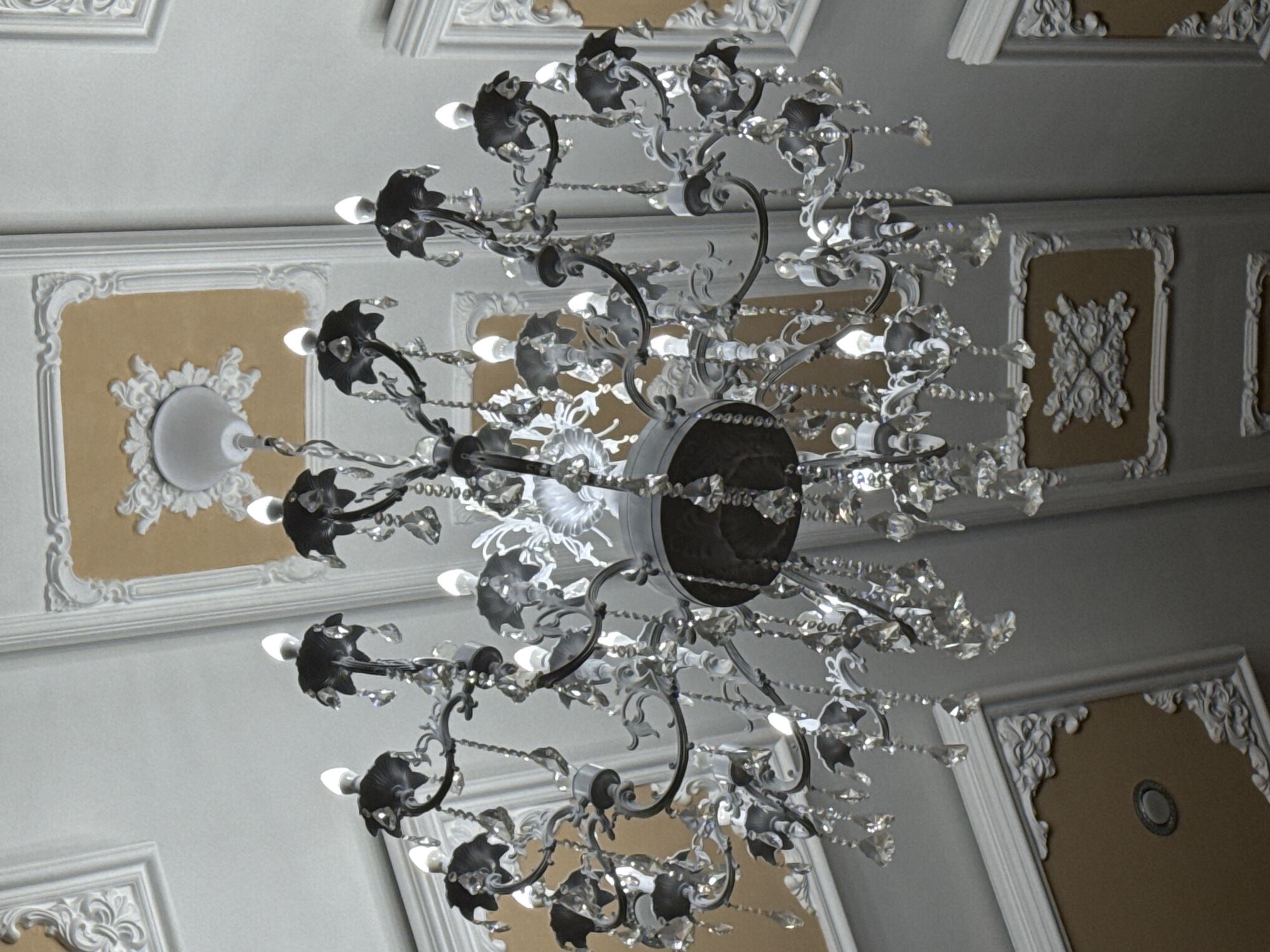 The image shows a view looking up at a chandelier hanging from a ceiling. The chandelier is ornate, with multiple tiers of decorative metal arms, crystal pendants, and lights. The ceiling features decorative molding and recessed panels with painted accents. The image location is likely to be inside a building with a high ceiling. The photograph was taken at an upward angle. The chandeliers lighting casts shadows on the ceiling. This location cannot be determined to be in the city of Votkinsk, Russia.