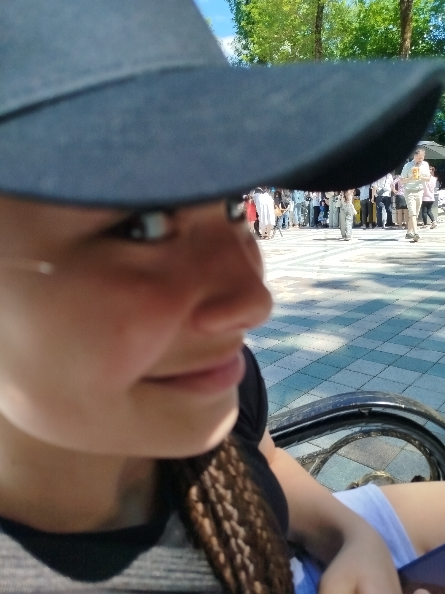 The image shows a close-up of a young woman wearing a dark baseball cap, looking to her right. She has a braid visible on her right side. In the background, a group of people are gathered in an outdoor public space with a checkered tiled ground. Some individuals are holding drinks, and there is a tent-like structure visible. The setting appears to be a sunny day with trees in the background. The scene is likely a public square or park in Stavropol, Russia, possibly during an event or gathering.