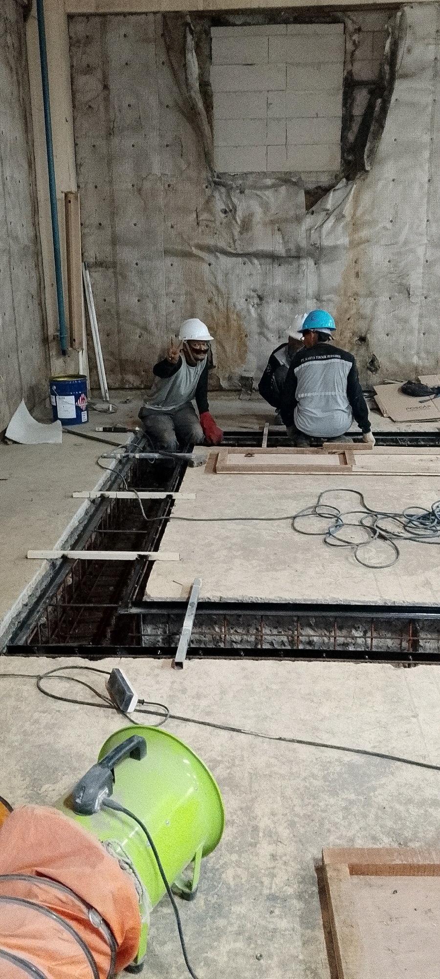 Two construction workers are observed in what appears to be an indoor construction site in Depok, Indonesia. One worker, wearing a white hard hat, grey shirt, and red gloves, is kneeling and gesturing with a peace sign towards the camera. The other worker, wearing a blue hard hat and a reflective vest with the text "PT. KARYA TEKNIK BERSAMA" on the back, is also kneeling and engaged in work. A third individual is partially visible behind them, also wearing a hard hat.
The environment is a concrete floor with a section cut out, revealing rebar and other construction materials below. Tools and equipment, including electrical cords, a green ventilation fan, and a blue paint can, are scattered around the work area. The walls of the space are concrete with visible insulation and some scaffolding-like structures. The lighting suggests an indoor setting with artificial illumination.