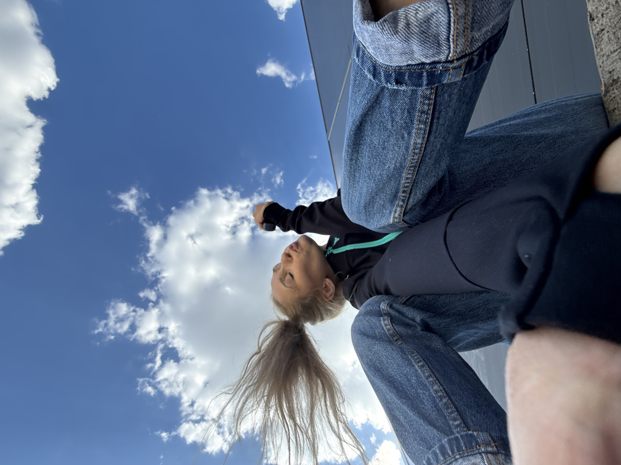 A low-angle shot captures a woman looking upwards at the sky. She has blonde hair pulled back in a ponytail, and is wearing a dark jacket with a light blue trim, and blue jeans. The background consists of a blue sky with white clouds. A gray wall is visible on the right side of the image. The location is Chaikovsky, Russia.