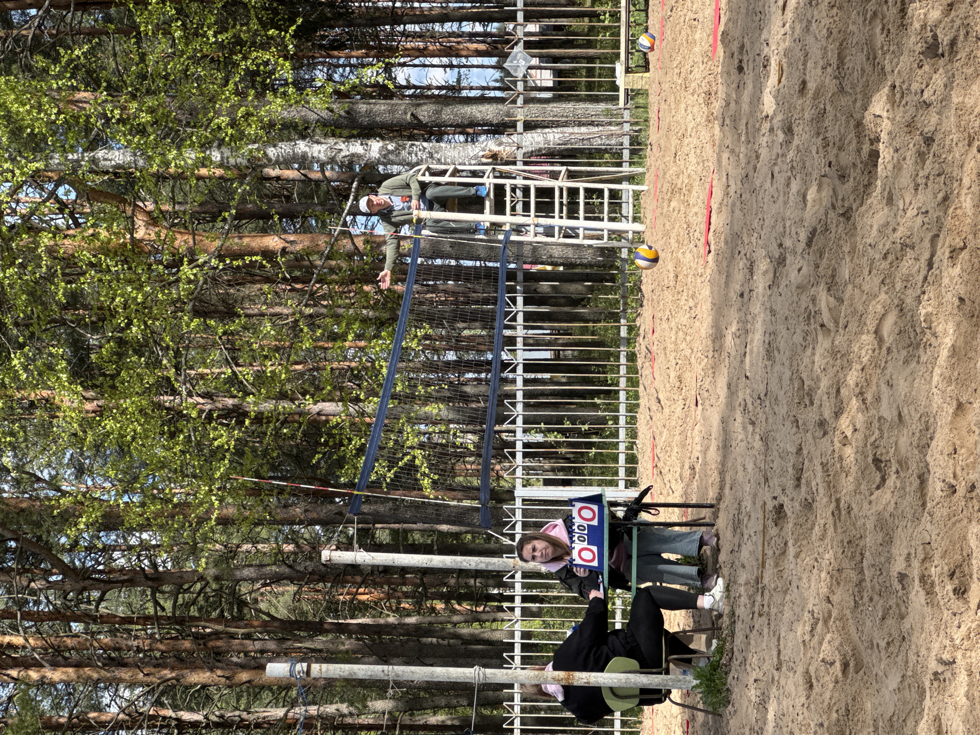 The scene shows a beach volleyball court in Yamyshevo, Russia. A referee stands on a high metal ladder above the net, extending one arm. A volleyball is suspended in the air near the net. Two individuals are seated at a scorer's table to the left of the net, one holding a manual scoreboard. The court surface is sand. The background consists of a dense forest. Another volleyball is seen in the distance near a barrier line.