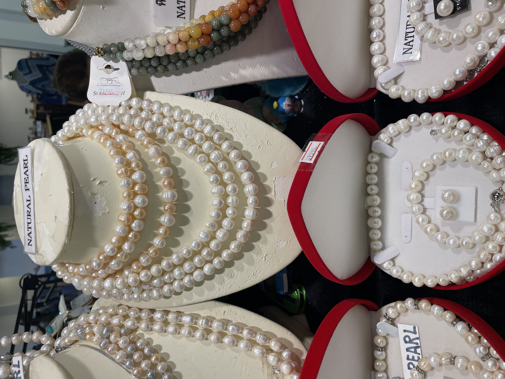 The image shows a display of pearl jewelry in what appears to be a retail setting, possibly a market stall or shop. There are several multi-strand pearl necklaces showcased on white mannequins. One necklace on the left is a mix of white and greyish pearls. The central display features a more voluminous necklace with pearls in shades of white and pale peach. To the right, a strand of multi-colored gemstone beads, possibly jade and agate, is visible.
In the foreground, three open red velvet jewelry boxes are displayed. The left box contains a pearl bracelet and earrings, with a label that says "PEARL". The middle box displays pearl earrings and a bracelet. The right box also contains a pearl bracelet and earrings, with a label that reads "NATURAL PEARL". Some of the jewelry pieces have price tags attached, with one clearly showing "฿1590.".
In the background, there are blurry hints of merchandise and mannequins, suggesting a larger retail environment. The overall impression is a commercial display of decorative items. The presence of a doll-like figure and what looks like a toy car in the background suggests it might be a shop catering to a diverse clientele, possibly even in a tourist-oriented area.
The request for the location in the city of Pattaya City, Thailand, cannot be fulfilled as there are no visible landmarks or contextual clues within the image to confirm this specific location. The image itself provides no geographical information.