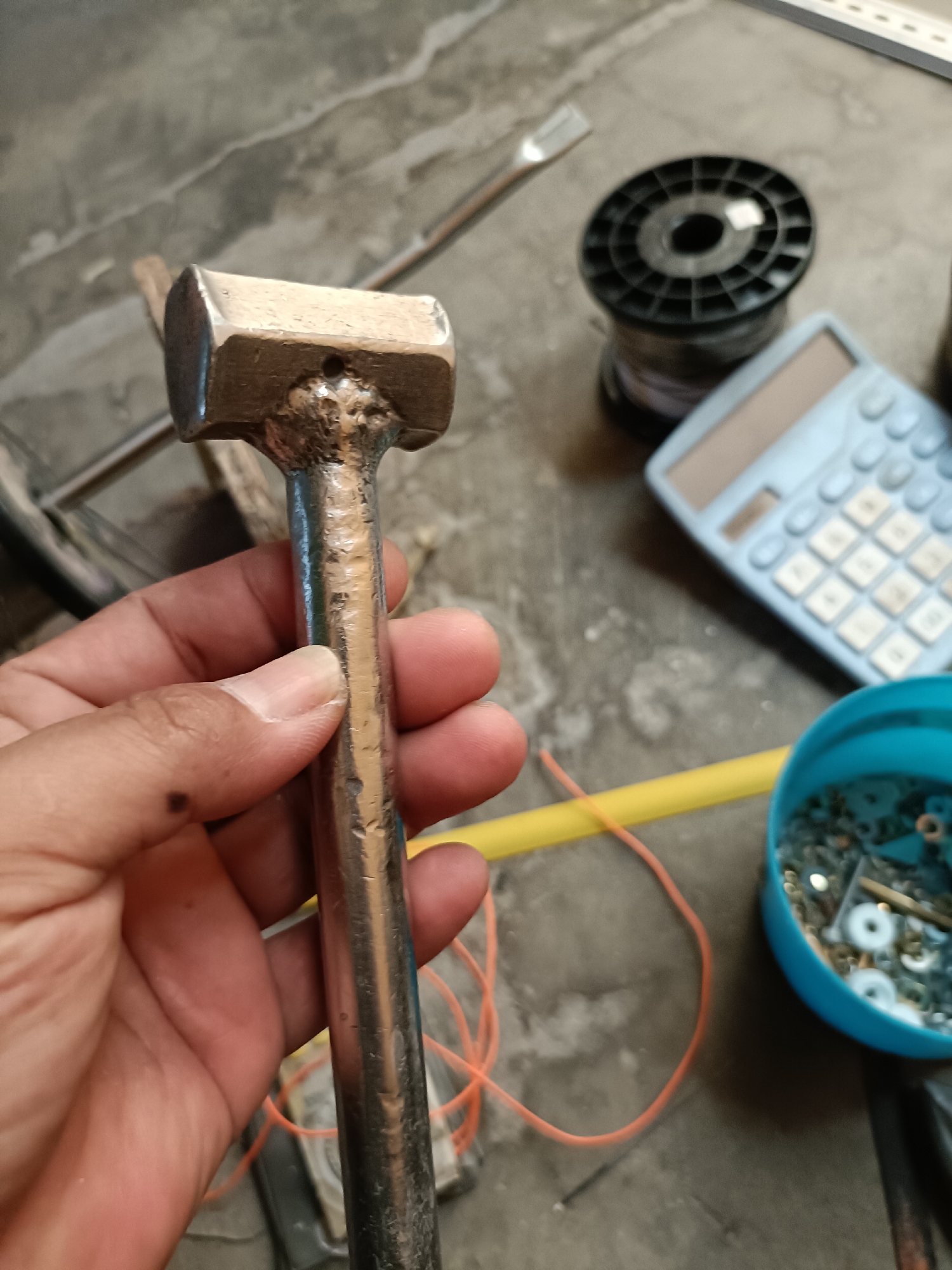 A close-up, high-angle shot shows a person's hand holding a small, metallic hammer with a rectangular head. The hammer appears to be handmade, with visible weld marks on the neck. The background suggests a workshop or storage area, with a concrete floor. Various objects are scattered around, including a blue plastic container filled with screws and washers, a yellow tube, an orange string, a spool of wire, a calculator, and what appears to be a screwdriver. The scene is dimly lit, with the focus on the hammer and the hand holding it. The image is taken in Telagamurni, Indonesia.