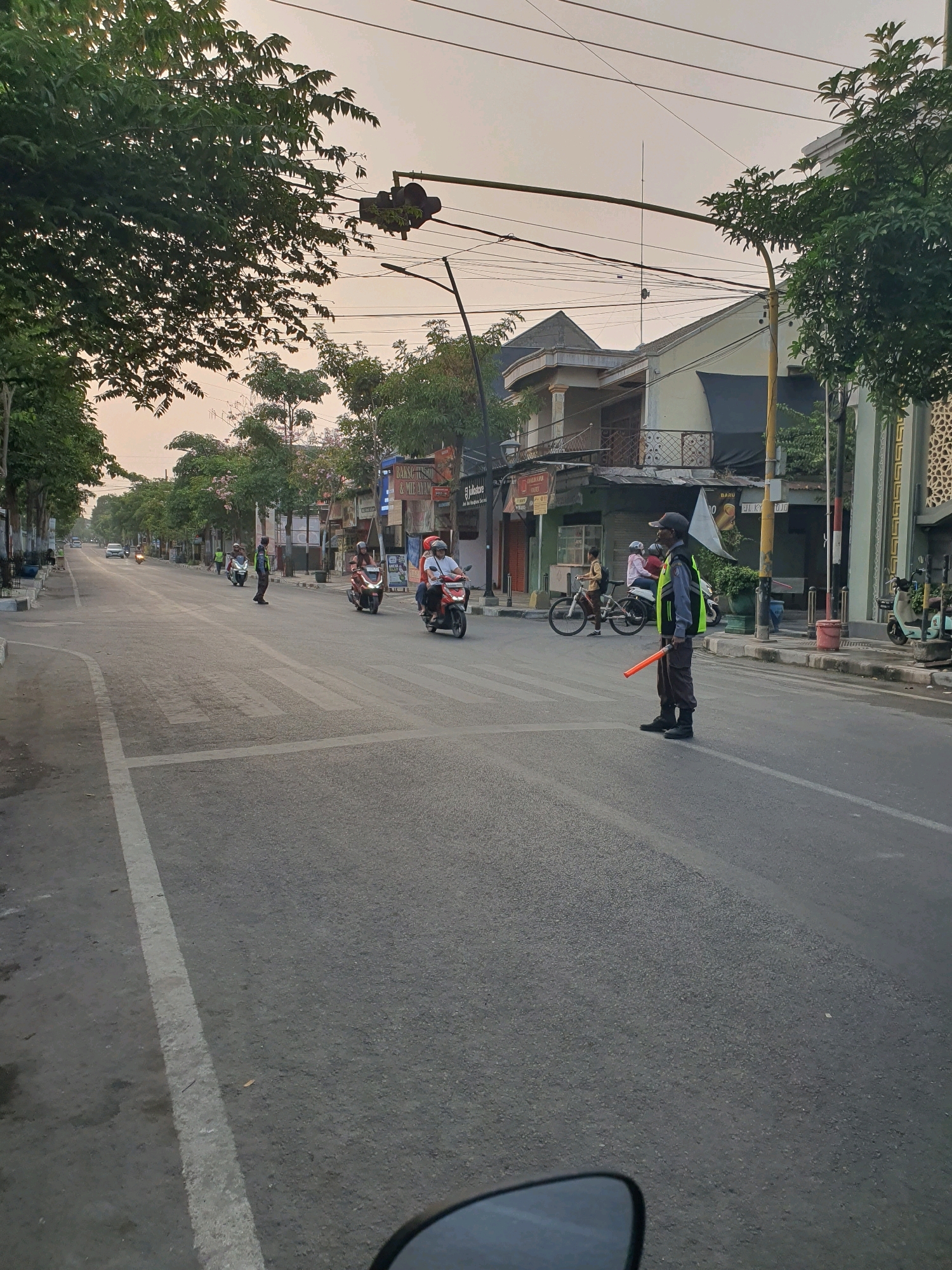 The image depicts a street scene in Bojonegoro, Indonesia, likely during the daytime given the ambient light. A traffic officer wearing a high-visibility yellow vest and a gray cap stands in the middle of the road, holding an orange traffic baton. The officer appears to be directing traffic. Several motorcycles are present on the road, with some riders wearing helmets. A bicycle is also visible, with a person riding it. Buildings line the street, some with awnings and storefronts. Trees are planted along the sidewalk, and power lines are strung overhead. The road surface is asphalt, with white lane markings and a pedestrian crossing. The sky is overcast, suggesting a diffused light source. The perspective suggests the photo was taken from a moving vehicle.