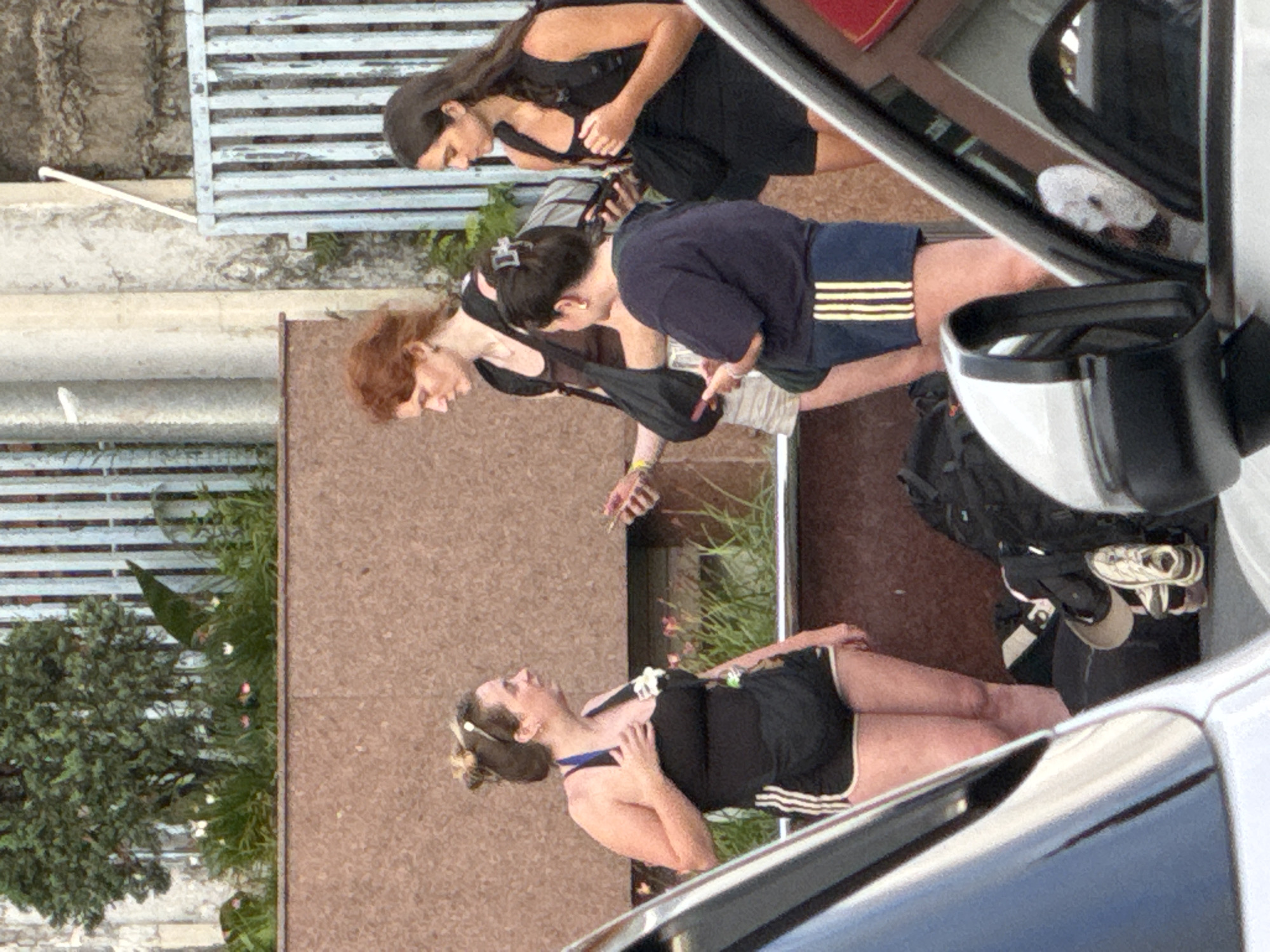The image captures an outdoor scene featuring three young women standing next to a building and a vehicle, likely in Da Lat, Vietnam, as suggested by the context of Da Lat. The woman on the left, with blonde hair tied up, wears a black tank top and black athletic shorts with white stripes. She holds a black strap in her hand. In the center, a woman with red hair and a brown tank top stands in front of a tall, textured brown wall and a light blue striped fence. She is wearing beige shorts. To the right, a woman with long dark hair and a black tank top stands near a similar fence. She is holding a phone. Behind the central and right figures, a dark backpack is visible. The foreground is dominated by a white vehicle, with its side mirror prominently displayed. The overall setting appears to be a street or sidewalk area with some greenery and urban elements.
