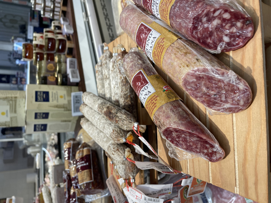 The scene depicts a detailed view of a deli counter or specialty food display within a retail establishment, likely a CONAD supermarket, located in Monsummano Terme, Italy. In the foreground, three plastic-wrapped salamis, labeled "SALAMINO TOSCANO" and displaying cut cross-sections of reddish, coarse-ground meat with white fat particles, rest on a light-colored wooden surface. Behind these, numerous whole, unwrapped dry-cured salamis with characteristic grayish-white casings and individual tags secured by red string are arranged on a wooden tiered display. Additional packaged cured meats are visible on the left side of the midground. The background features shelving units; on the right, multiple small jars of preserves or honey are stacked on miniature wooden pallets, while in the center, several tall, rectangular cardboard boxes branded "SAPORI D'TOSCANA" are stacked. A digital display showing "26.xx" is present on a lower shelf. The environment is brightly lit, consistent with an indoor commercial setting, and no individuals are visible.