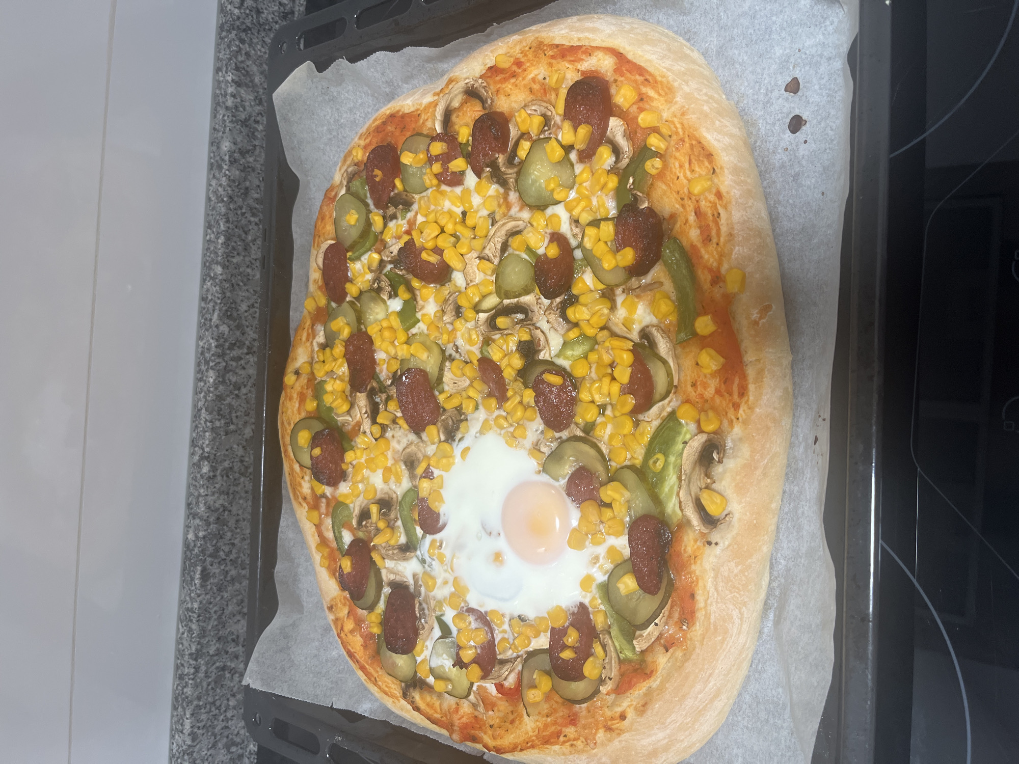 An overhead shot depicts a freshly baked pizza on a baking sheet lined with parchment paper. The pizza is topped with tomato sauce, corn, mushrooms, sliced pickles, sausage slices, green pepper strips, and a fried egg in the center. The crust is thick and slightly browned. The pizza is placed on a baking sheet, which is sitting on a stovetop or countertop. The image appears to have been taken indoors in a kitchen. It cannot be confirmed that this is Mogán, Spain.