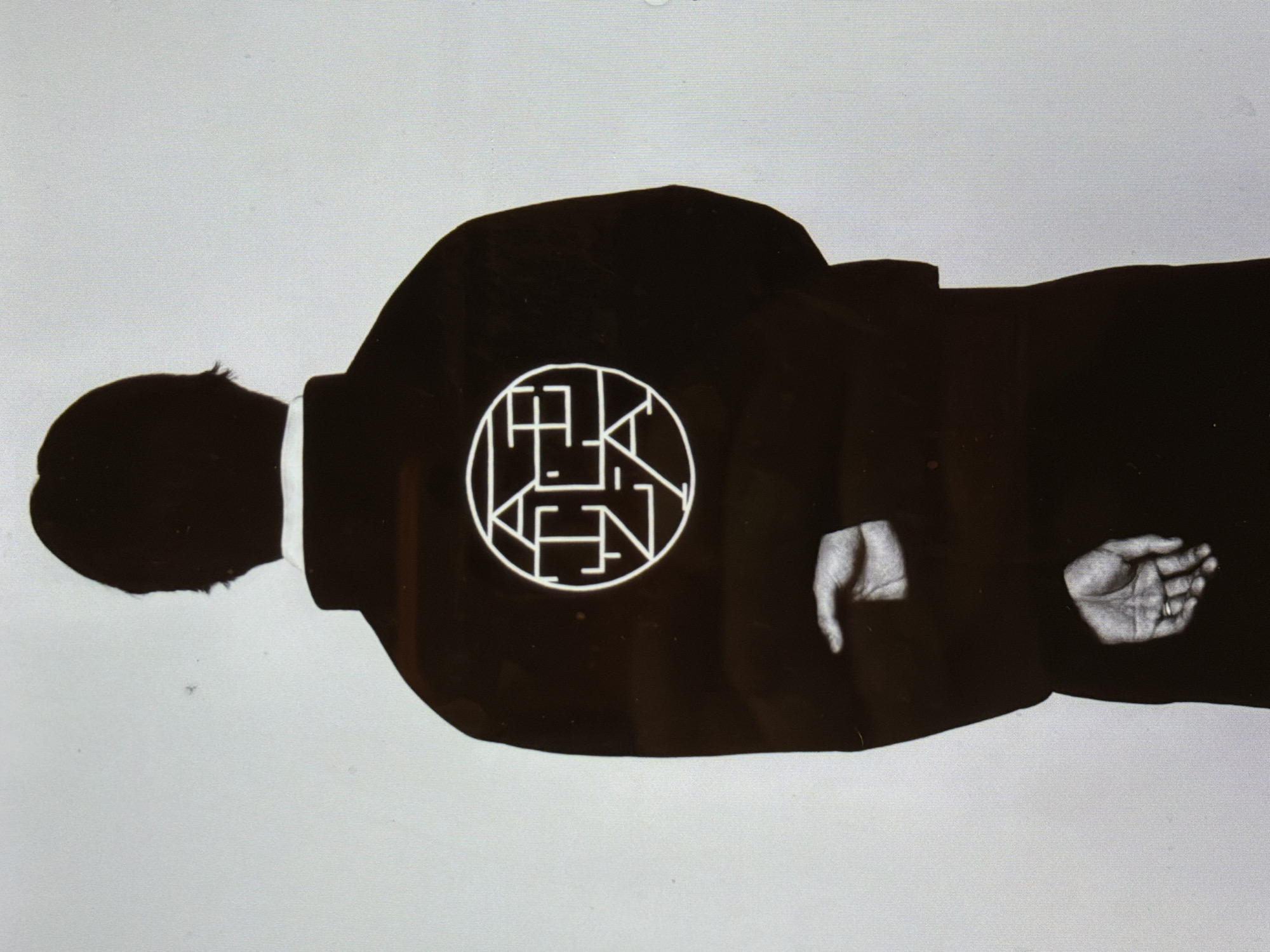 The image depicts a silhouette of a man in a suit. He is seen from the side with his back to the viewer. A white circular logo is imprinted on the back of his jacket. His hands are visible, appearing clasped in front of him. The background is plain white. The setting is not identifiable as New York City.
