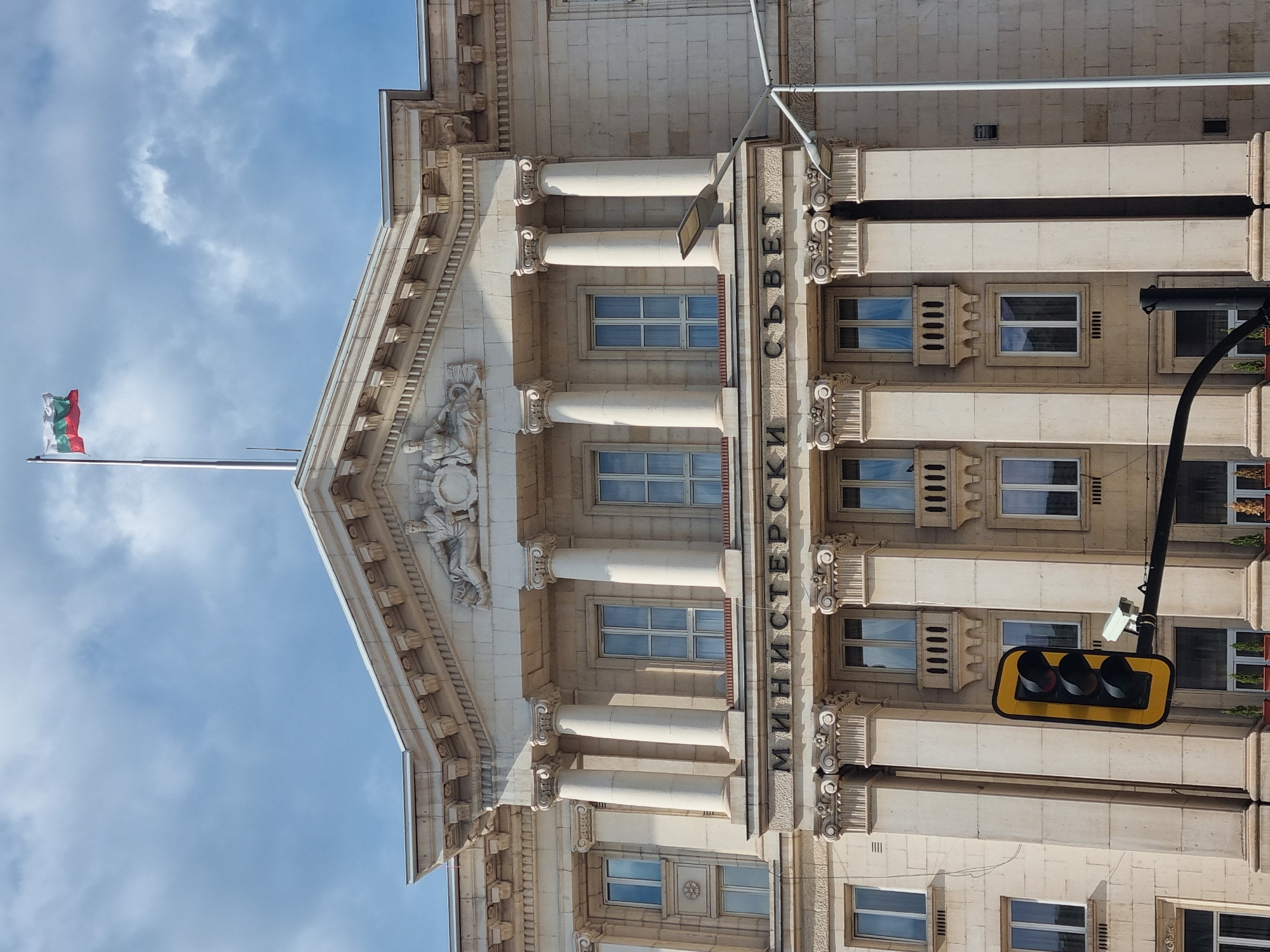 The image captures a section of a large, ornate building in Sofia, Bulgaria. The building, likely a government ministry, is constructed from light-colored stone, with classical architectural elements such as columns, decorative moldings, and sculptures. The Bulgarian flag flies atop the building. The Cyrillic text "МИНИСТЕРСКИ СЪВЕТ" ("Council of Ministers") is prominently displayed on the facade. A modern traffic light fixture is attached to the side of the building. The sky is blue with some clouds.