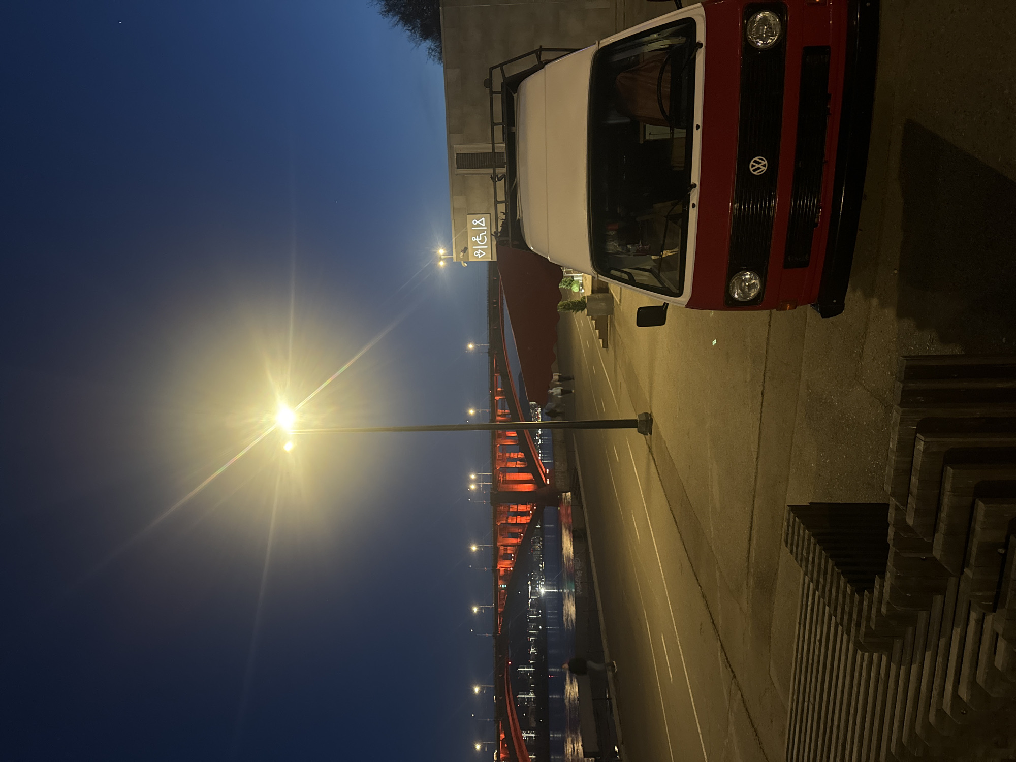 The image shows a red and white Volkswagen van parked on a paved surface next to a bridge in Krasnoyarsk, Russia. A streetlight illuminates the scene, and a sign with wheelchair and restroom symbols is visible near the van. The bridge, with red supports, spans a body of water reflecting city lights. A stacked wooden structure is present on the right side of the frame. The sky is dark blue.
