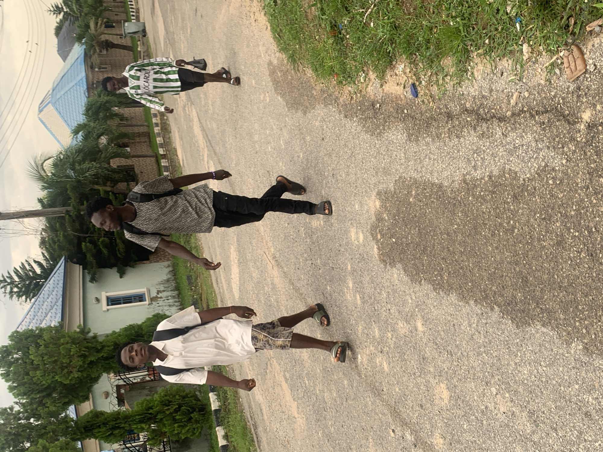 Three young men are walking on a paved road in an urban environment. The man on the left is wearing a white polo shirt, patterned shorts, and dark sandals. He has a black backpack on. The middle man is wearing a black and grey patterned t-shirt, black pants, and dark sandals, also with a black backpack. The man on the right is wearing a green and white striped long-sleeved shirt with the word "IOCAROL" visible, black shorts, and dark sandals. He is carrying a black plastic bag. In the background, there are houses with distinctive blue roofs, some palm trees, and manicured bushes. The road is asphalt with visible tire marks and some scattered debris. The sky is overcast. The scene appears to be in a residential area of Abuja, Nigeria.