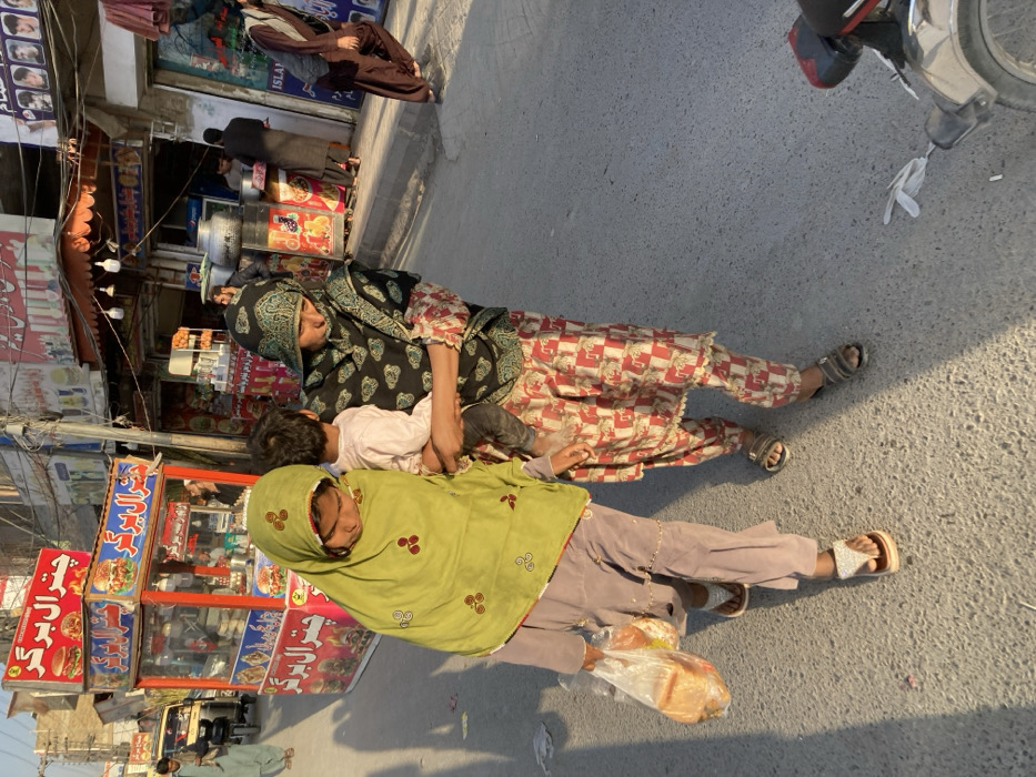 The image portrays an urban street scene in Quetta City Tehsil, Pakistan, characterized by natural sunlight creating long shadows. Three individuals are prominently featured in the foreground, walking along an asphalt road. On the left, a female wearing a light green headscarf and light-colored shalwar kameez carries a clear plastic bag containing loaves of bread in her left hand. Beside her, an adult female, dressed in a dark patterned headscarf and a red and white patterned shalwar kameez, carries a small child, clothed in a light top and dark trousers, on her left hip. Both women's heads are slightly bowed.
The background shows a busy commercial street lined with various shops and food stalls. A red-framed glass food cart, adorned with "Chitral Burger" in Urdu and burger illustrations, is positioned in the mid-ground. Behind it, a yellow auto-rickshaw is partially visible. Further right, additional shops display signs in Urdu, some featuring images, and a stall with a red canopy is present. Several indistinct figures are visible in the background, walking or standing near the establishments, including a man in light green on the far left and another in brown on the right. A utility pole with overhead wires rises on the right side of the street. In the bottom right corner, the rear wheel, fender, and exhaust pipe of a grey motorcycle or scooter are visible, with some white debris on the road nearby.