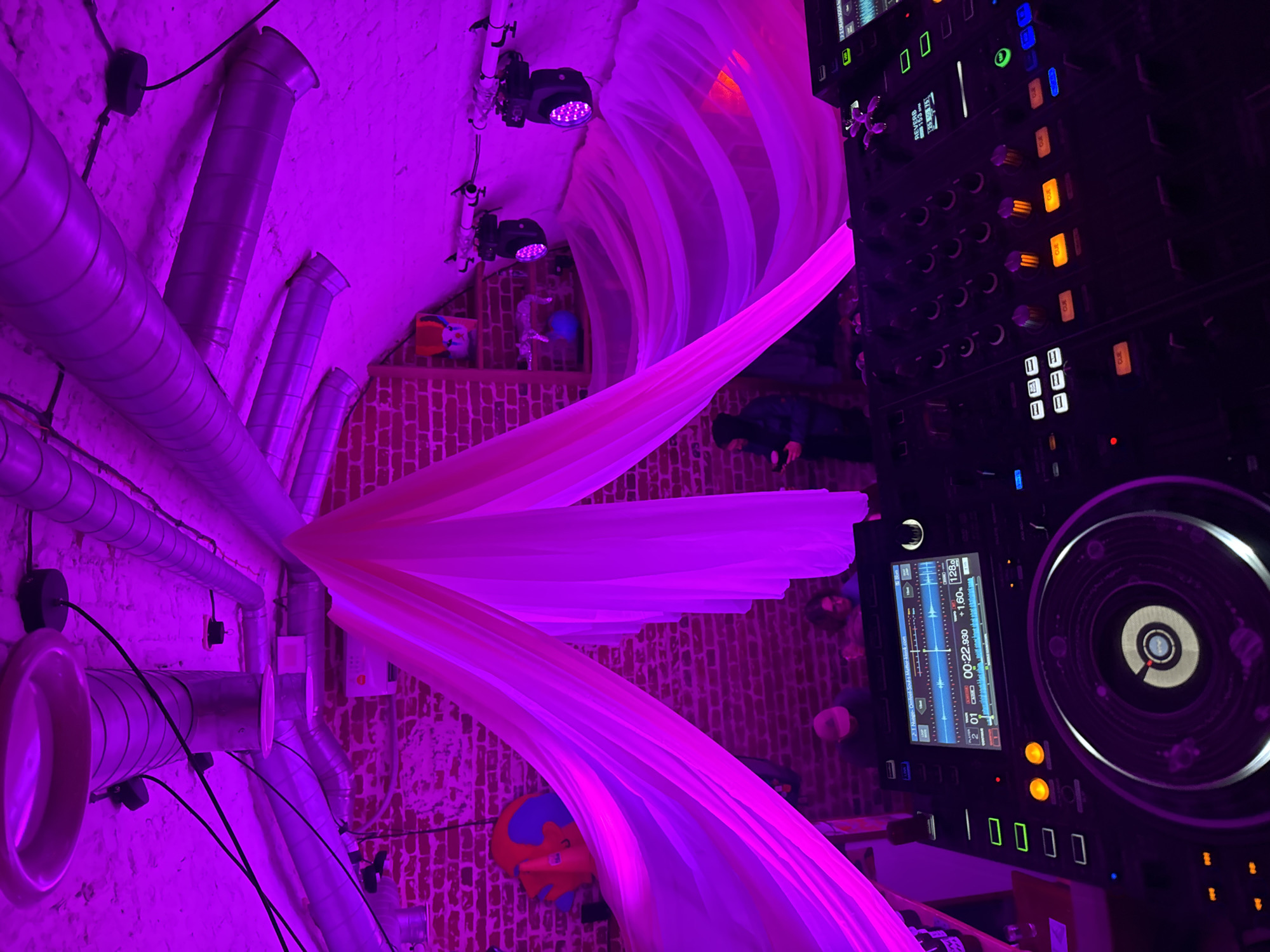 This indoor scene, possibly in a club or event space in Krasnoyarsk, Russia, is lit with bright, primarily purple lighting. The walls are a combination of exposed brick and metallic ventilation ducts. Fabric drapery is suspended from the ceiling, adding to the ambiance. Overhead, stage lights are visible.
On the right side of the frame, a DJ mixing board with screens and various buttons is prominently displayed. Several people are vaguely visible in the background, their activities unclear. A child's toy or object is also present.