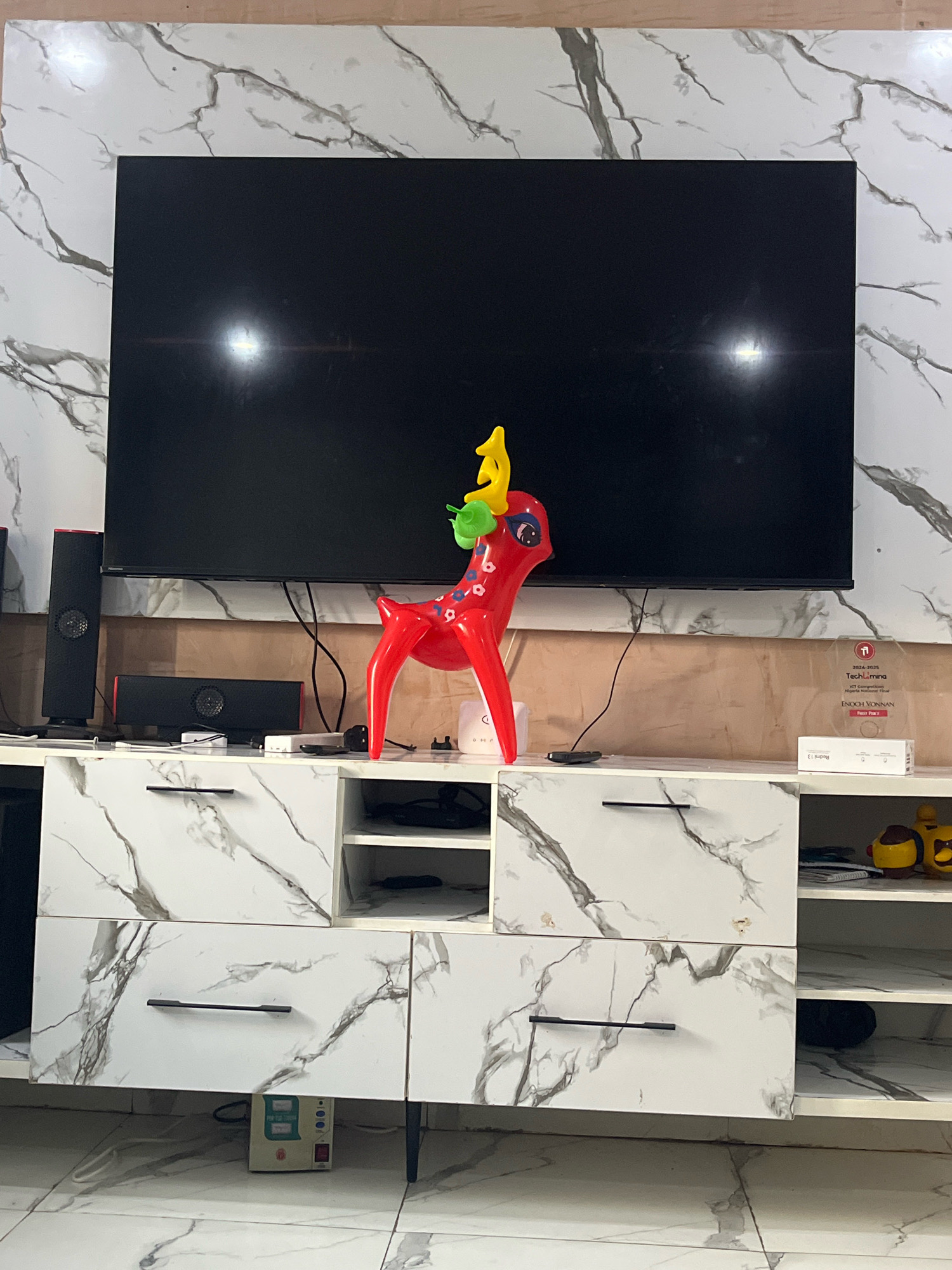 The image shows a living room in an apartment, possibly in Jos, Nigeria. A large black television is mounted on a wall with a marble-patterned finish. Below the television is a white entertainment center with drawers and shelves, also featuring a marble pattern. On top of the entertainment center sits a red decorative reindeer figure with yellow antlers and a green leafy detail on its head. To the left of the television are two black speakers. To the right of the reindeer is a clear acrylic award with text and logos, and a white box labeled "Redmi 13". On a shelf of the entertainment center, a yellow toy character can be seen. A small beige electrical device with buttons and a red switch is placed on the floor beneath one of the drawers. The floor also has a marble tile pattern. The overall impression is a modern and tidy living space.