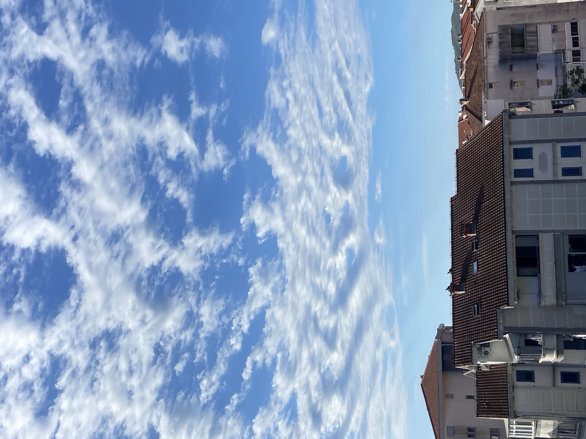 The image shows a view of buildings in Budva, Montenegro, with a bright blue sky filled with white, wispy clouds. The buildings are a mix of styles, with some having terracotta tile roofs and others having flat roofs. The buildings appear to be multi-story residential or apartment buildings. There are visible windows, balconies, and air conditioning units on the facades. The foreground is dominated by the rooftops of several buildings, with the sky occupying the upper half of the frame. The scene suggests a sunny day in an urban environment.
