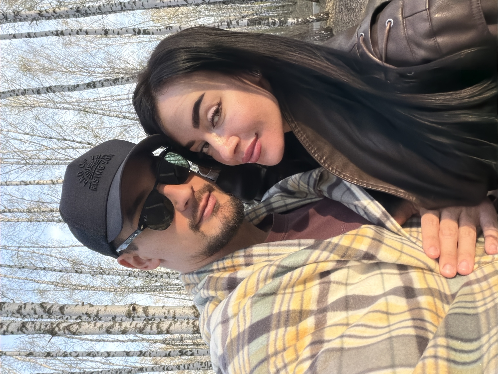 A couple poses for a photo in front of a birch tree forest, possibly in the vicinity of the city of Krasnoyarsk, Russia. The man wears a dark baseball cap and sunglasses, and a yellow and grey plaid jacket is draped over his body. The woman has dark hair and is wearing a brown leather jacket. She is leaning her head against his. The background consists of numerous birch trees with white trunks and bare branches. The sky is visible between the branches.