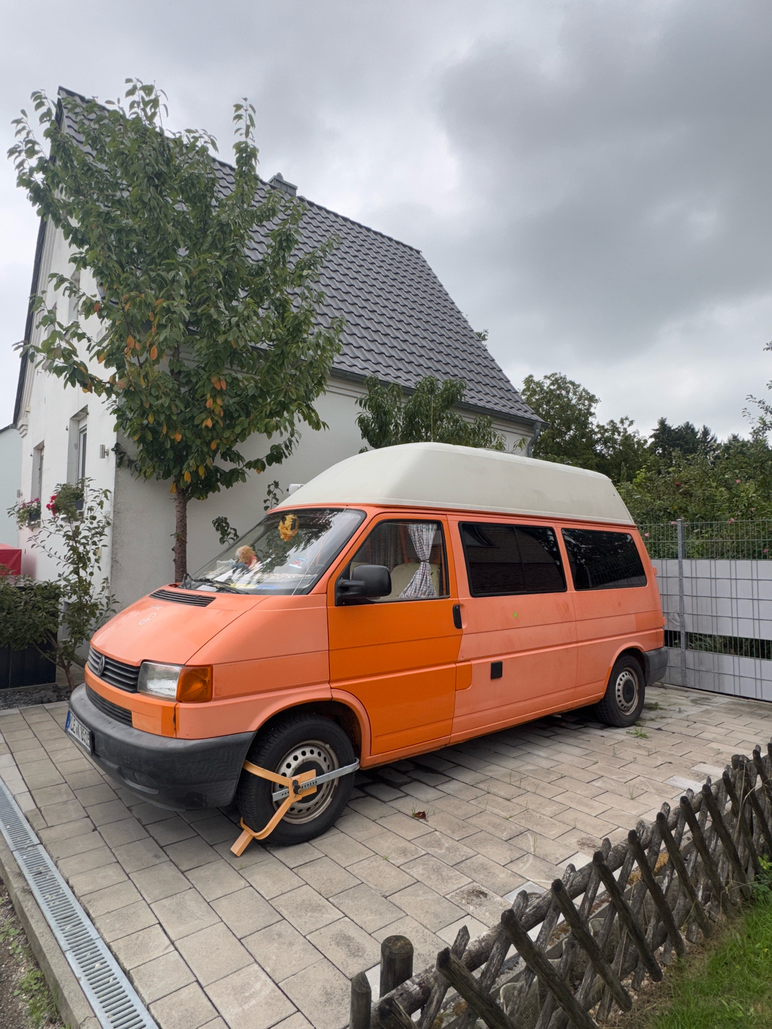 In Cleves, Germany, an orange camper van with a pop-top roof is parked on a paved driveway in front of a white house with a grey tiled roof. A tree with green and yellow leaves stands to the left of the van, near the house. The van has a wheel clamp attached to its front wheel. A metal fence is visible behind the van, and a wooden fence runs along the right side of the driveway. The sky is overcast.