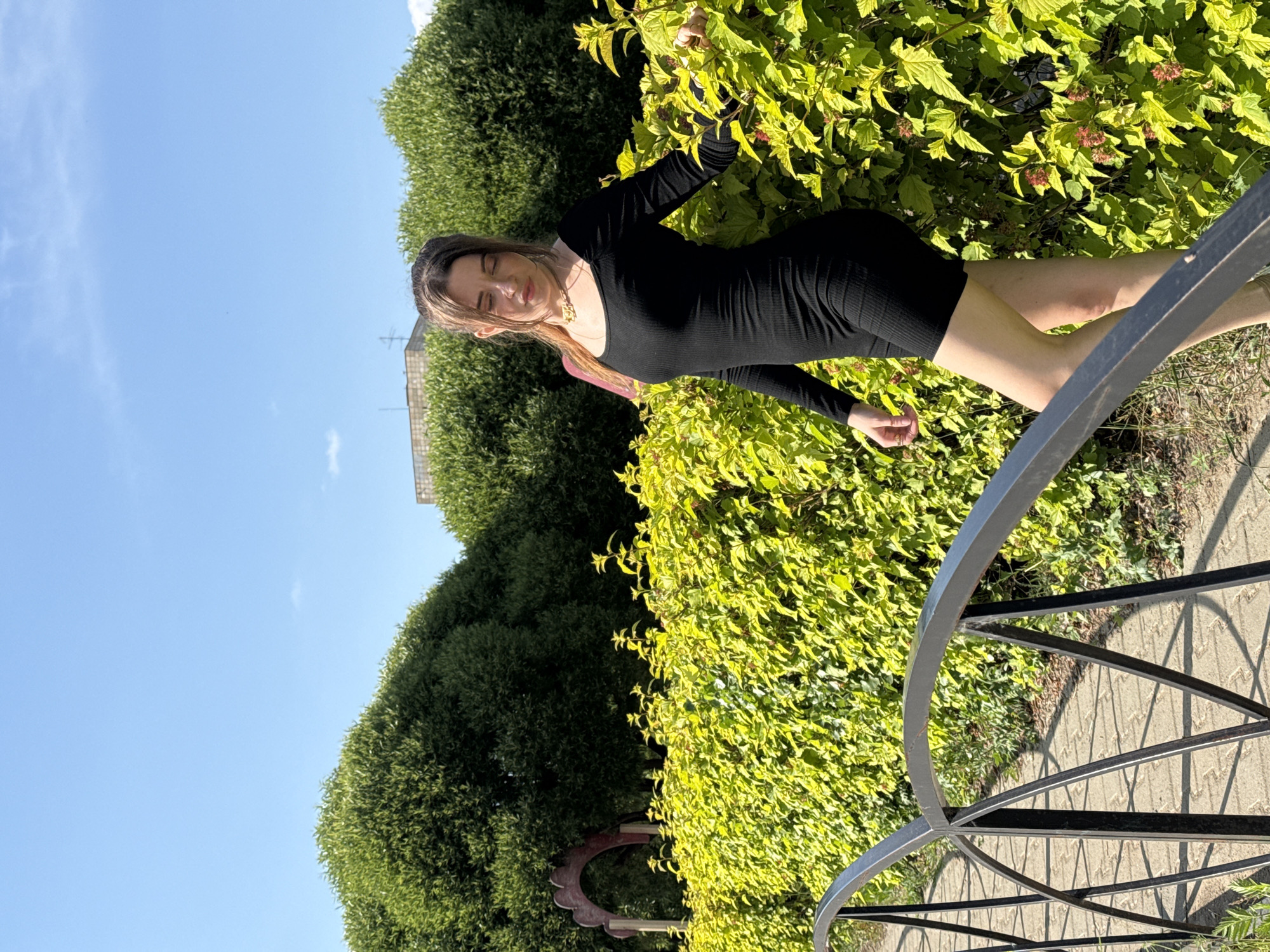 A young woman with long brown hair, wearing a form-fitting black dress and a gold necklace, stands on a paved pathway next to a dense green hedge. She is leaning her right arm on the hedge, with her head tilted down and eyes closed. A black metal railing, part of a small bridge or walkway, runs diagonally across the foreground of the image. Behind the hedge are several large, rounded green bushes and a glimpse of a building in the distance, all under a clear blue sky.