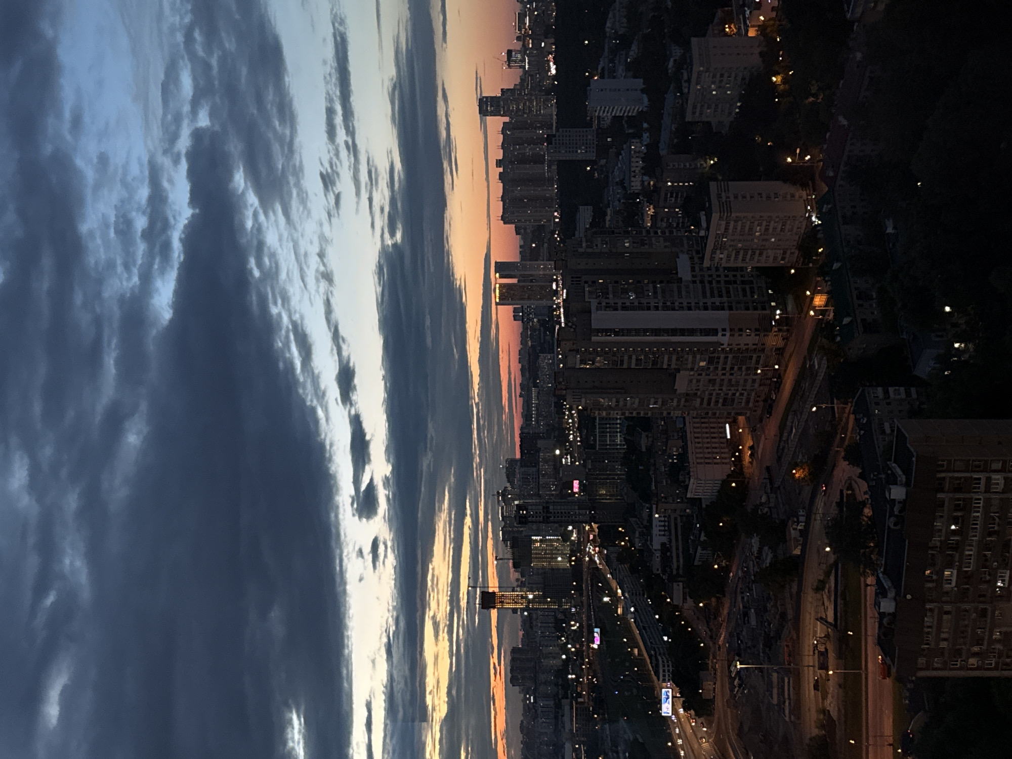 This is a vertical, wide-angle shot of an urban landscape during twilight. The sky on the left is filled with dramatic, dark gray clouds streaked with lighter tones, suggesting a turbulent or overcast sky. To the right, the sky transitions through hues of soft orange and pink, indicative of sunset or sunrise.
Below the sky, a densely populated city skyline stretches across the frame. Numerous tall buildings, appearing to be apartment blocks and office structures, are visible. Many of these buildings are illuminated, with lights shining from windows, suggesting that night is falling or has begun. The city lights create a warm glow against the darkening sky. Roads and what appear to be some green spaces or parks are interspersed between the buildings. A few vehicles can be seen on the roads, their headlights and taillights creating streaks of light.
The scene is set in Khimki, Russia, as indicated by the location context. The time of day is clearly twilight, with the transition from daylight to night being a prominent feature. The weather appears to be changing, with dramatic clouds in the sky. The overall emotion evoked by the image is one of transitioning light and the bustling activity of a city as day ends. There is no discernible text within the image.