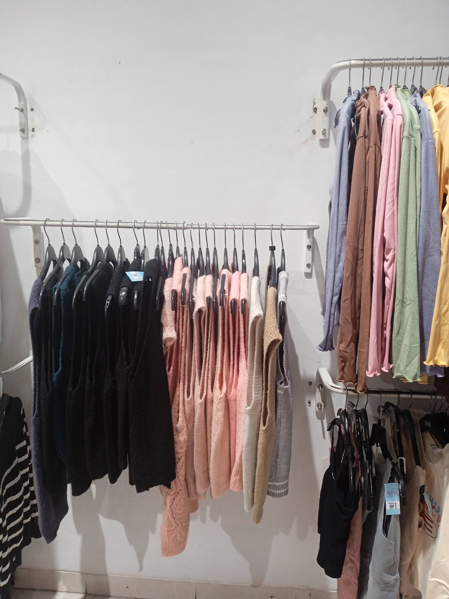 The image depicts a retail display of clothing on metal racks against a white wall. On the upper rack, a row of colorful garments, including pink, green, blue, and yellow tops, are neatly hung. The lower rack features a variety of sleeveless tops and sweaters in black, dark blue, and pink hues. The garments appear to be new and are presented for sale. The scene suggests a clothing store or boutique setting. No people are visible in the image. The location is identified as Semarang, Indonesia, though no specific landmarks or indicators of this location are present in the photograph.