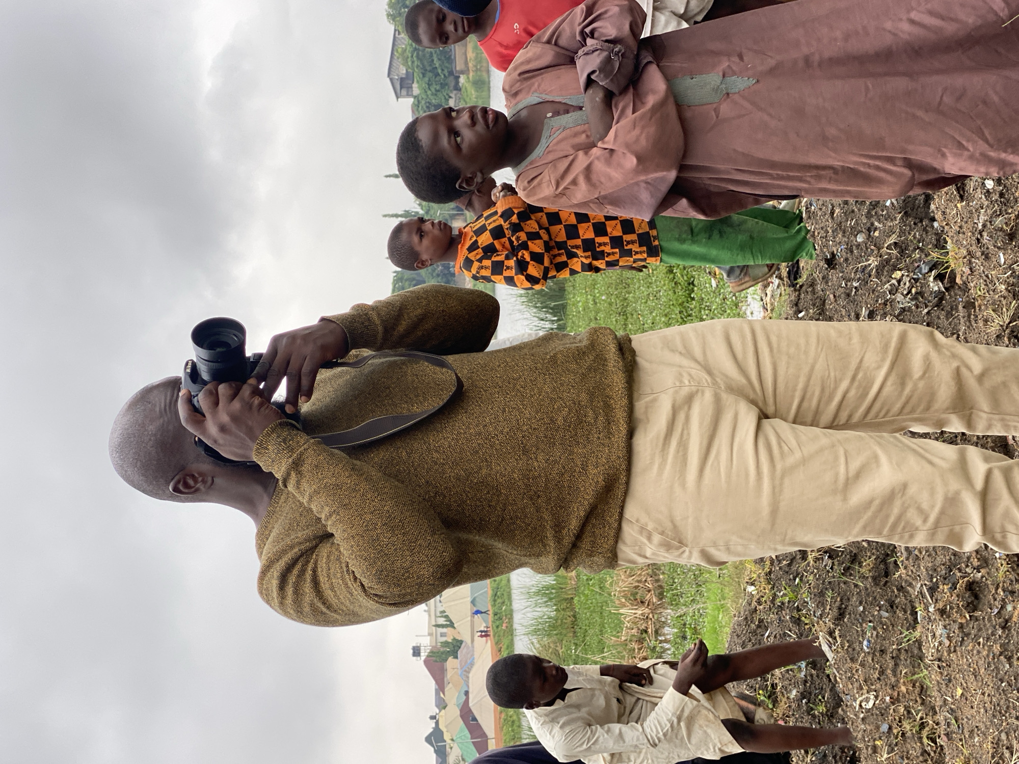 The image depicts an outdoor scene in the city of Kawo, Nigeria, with a cloudy sky overhead. In the foreground, a bald Black man is holding a black Nikon camera to his face, taking a photograph. He is wearing a textured, mustard-yellow sweater and light brown pants. To his left, a young Black boy in a light-colored tunic is crouched down, seemingly examining something in the dirt. Further back, several other Black children are visible. One boy in the middle wears an orange and black checkered shirt over green pants, and another to his right wears a red shirt. Behind the children and the photographer, a body of water with green reeds can be seen, and in the distance, there are colorful tents or temporary structures and buildings. The ground in the foreground is uneven and appears to be made of dark soil mixed with debris.