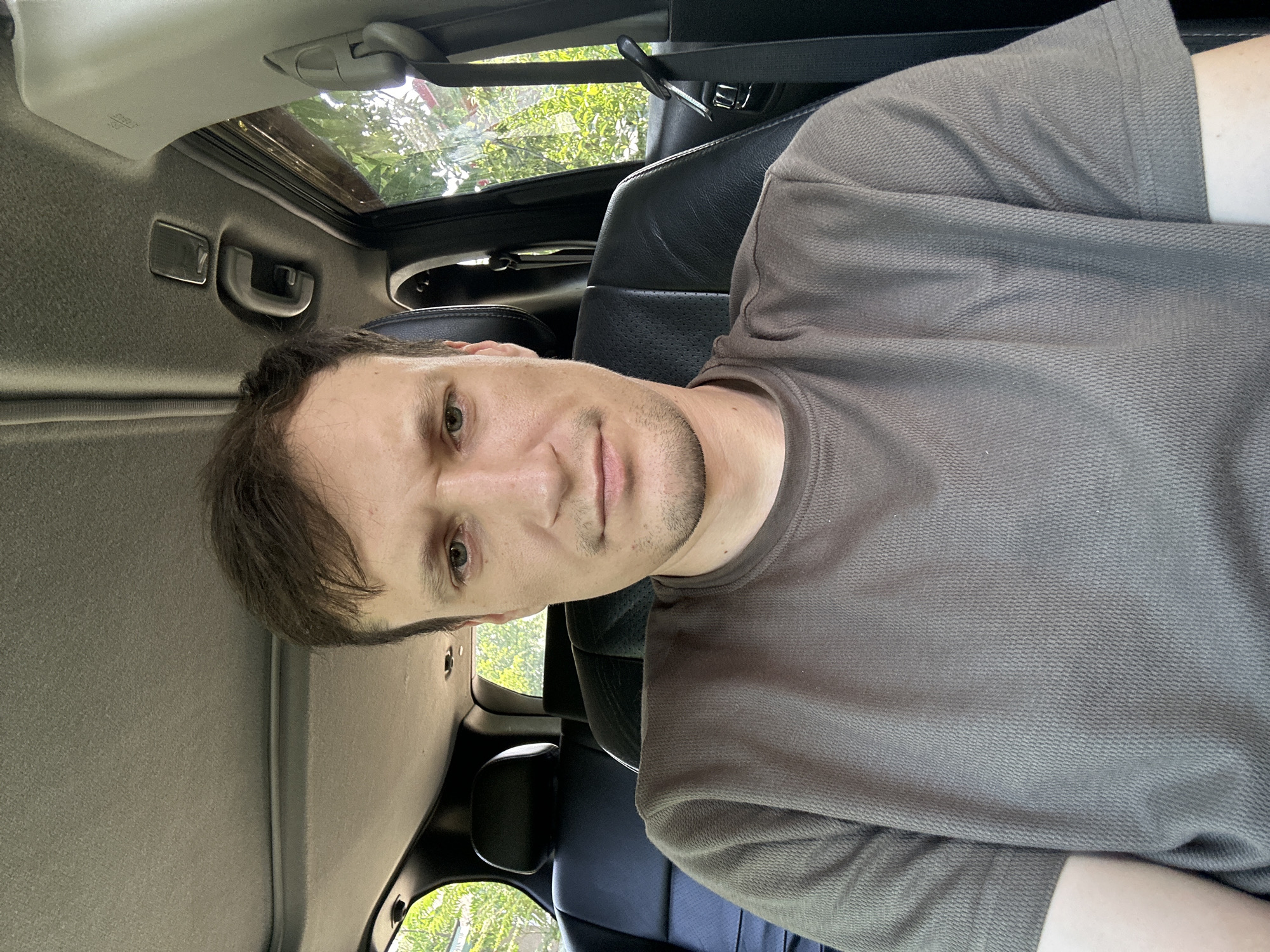 A medium shot shows a man sitting in the driver's seat of a car, looking directly at the camera. He has brown hair, light skin, and is wearing a gray t-shirt. The interior of the car is visible, with black leather seats and a sunroof. Trees and foliage can be seen through the car windows, suggesting an outdoor location, possibly in the city of Makiv, Ukraine, though this cannot be definitively confirmed. The overall lighting suggests it is daytime.