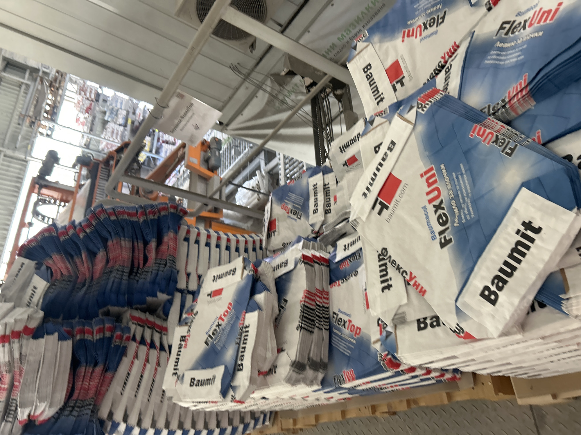The image shows a warehouse in Elin Pelin, Bulgaria, filled with stacks of construction material bags. The bags are predominantly white, blue, and red, featuring the brand name "Baumit" and product names like "FlexTop" and "FlexUni". Several bags are partially open or creased, suggesting they are either recently used or in storage. In the background, industrial machinery and shelving units are visible, indicating an active commercial or industrial setting. The lighting appears to be artificial, typical of an indoor warehouse environment. There are no people clearly visible in the frame.