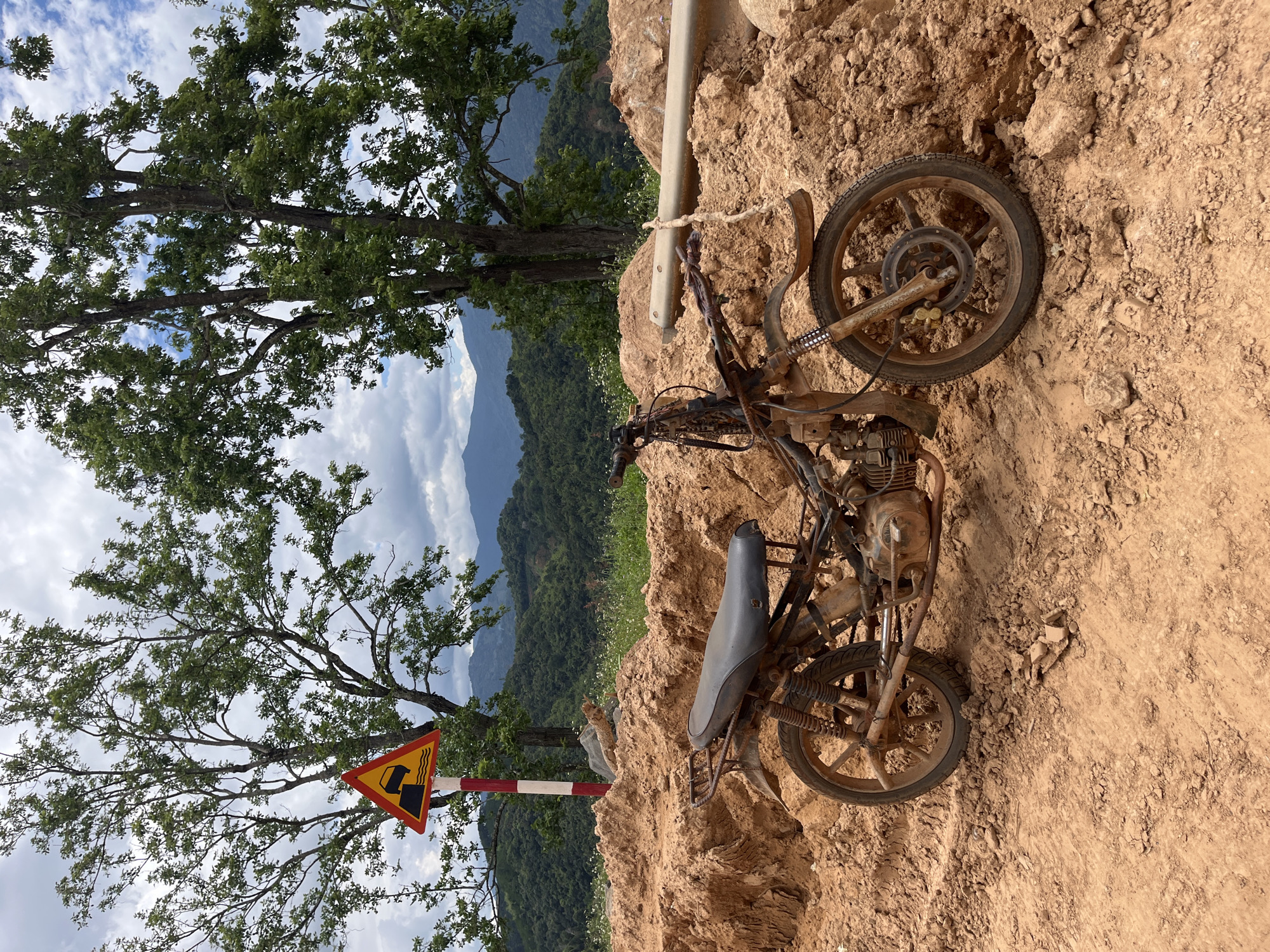 A rusty, dilapidated motorcycle sits abandoned on a dirt pile in a rural, mountainous landscape. The motorcycle is incomplete, with its front fairing, handlebars, and side panels missing. Its tires are worn, and the frame is coated in a thick layer of reddish-brown dirt. The surrounding environment is characterized by a steep, uneven terrain with large mounds of earth, possibly from construction or excavation. In the background, lush green hills roll into the distance under a cloudy sky. A yellow triangular road sign with a black symbol depicting a sharp descent or drop-off is visible to the left of the motorcycle, mounted on a striped red and white pole. The scene suggests a remote location in Nao Sẻ, Vietnam, likely a construction site or an area undergoing development, where the motorcycle has been left behind due to its condition or the challenging terrain. No people are visible in the image.