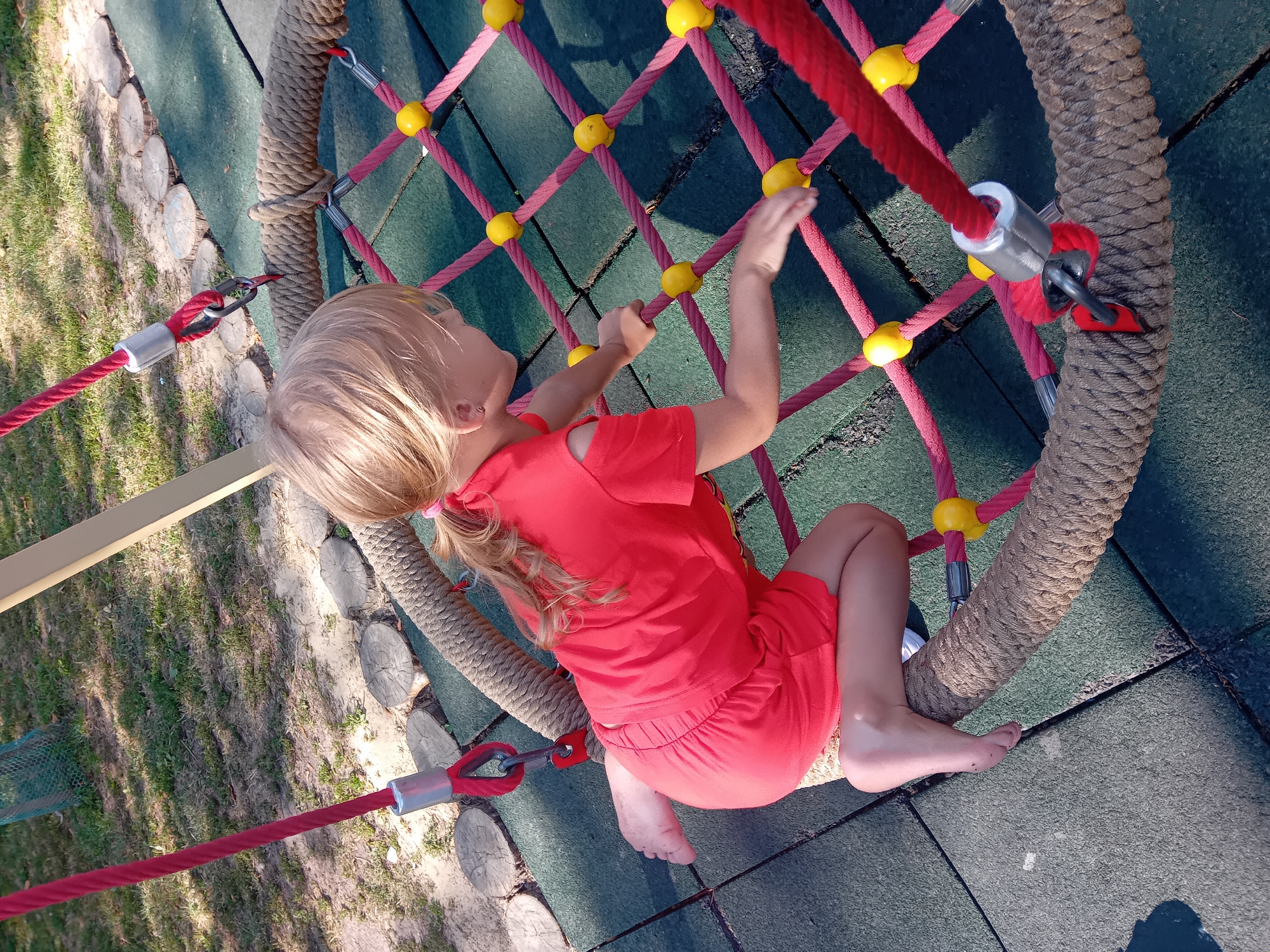 The image shows a young girl with blonde hair in a red outfit playing on a rope net swing in a park. The swing has thick rope edges and a web of thinner red ropes with yellow connectors. The girl is kneeling on the edge of the swing, facing away from the camera, with her hands on the rope net. Her left leg is bent, and her foot is visible. The ground beneath the swing appears to be made of dark green rubber matting, with some grassy areas and logs visible in the background. The scene suggests an outdoor playground environment, likely in Chuhuiv, Ukraine, given the context.