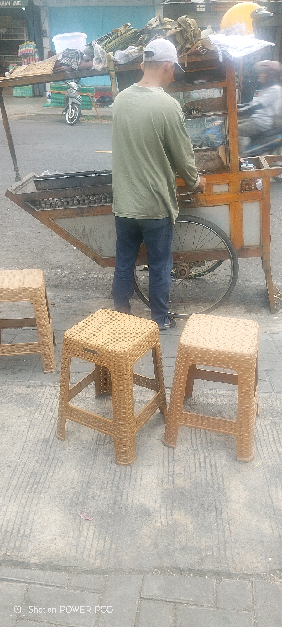 In an outdoor urban environment, a man wearing a light green long-sleeved shirt, dark blue pants, and a white baseball cap stands behind a food cart. The cart, made of wood and metal, has a large wheel at its base and is laden with what appears to be food items and packaging. In the foreground, three brown plastic stools are arranged on a patterned concrete surface. A motorcycle is visible in the background, parked next to a building with blue siding. A yellow helmet is perched on top of the food cart. The overall scene suggests a street food vendor setting up or operating their business.