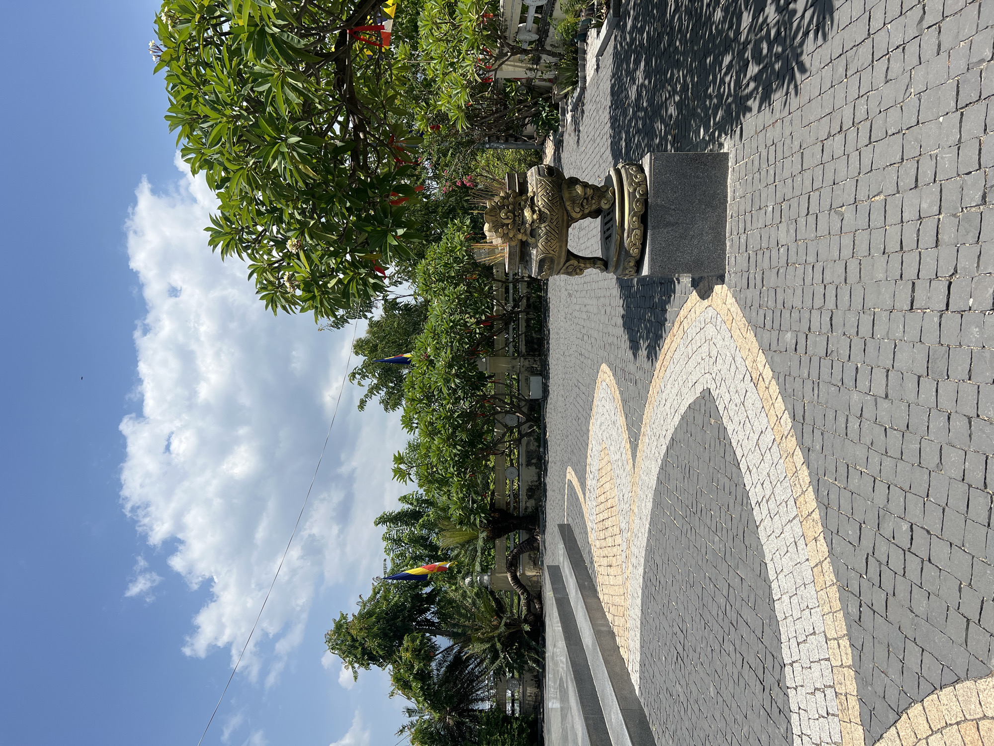 An outdoor shot captures a paved courtyard with a decorative serpentine pattern made of yellow and white stones against a dark gray cobblestone ground. To the right, a large, ornate bronze incense burner with dragon motifs sits on a dark stone pedestal, filled with incense sticks. Lush green trees with scattered red and yellow ribbons hanging from their branches line the background. In the middle ground, a white stone wall with decorative elements runs behind a line of trees and plants. To the left, a set of wide, gray stone steps leads up to a raised area. Flags in various colors, including red, yellow, and blue, are visible fluttering in the distance. The sky above is a bright blue, dotted with fluffy white clouds. The scene appears to be a serene courtyard, possibly at a temple or religious site.