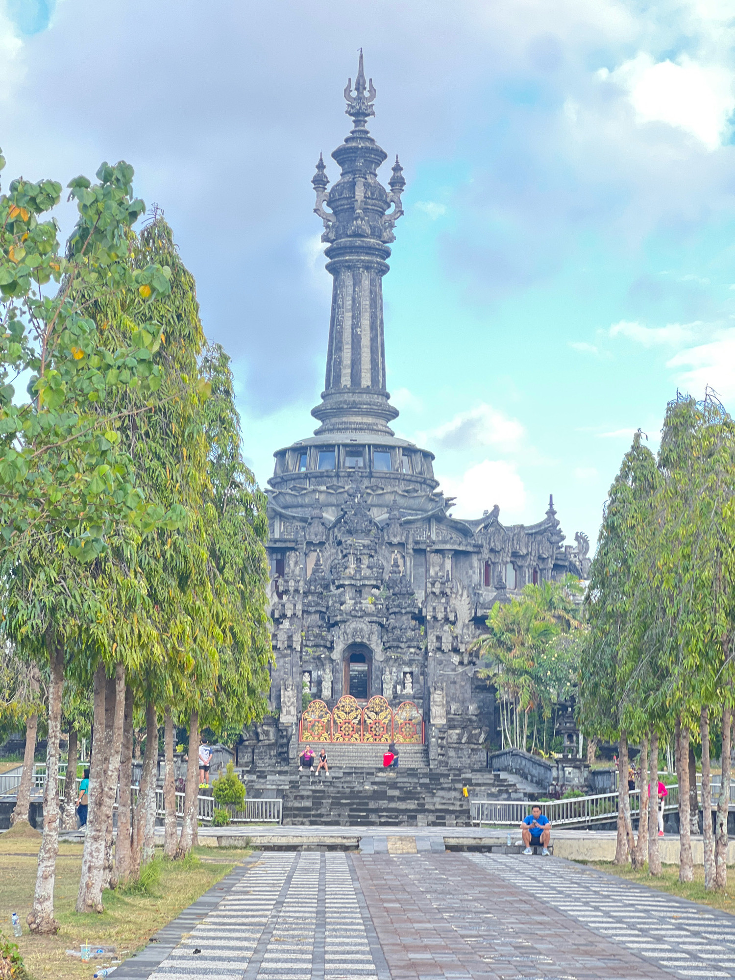 The image shows the Bajra Sandhi Monument in Denpasar, Bali, Indonesia. The monument, a tall, ornate stone structure, dominates the center of the frame. It is surrounded by lush green trees and a paved walkway. In the foreground, a man in a blue shirt and shorts is sitting on a ledge. Further back, several people are visible on the steps leading up to the monument. The sky is partly cloudy.