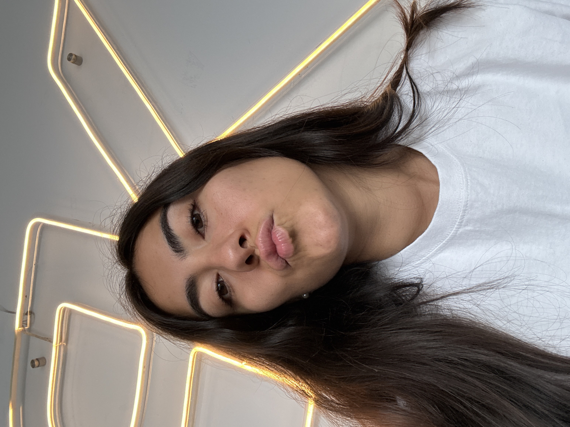 A young woman with long, dark hair is pictured in a selfie. She is wearing a white t-shirt. She is making a kissing face. Behind her is a wall with a neon light fixture. The location cannot be determined from the image.