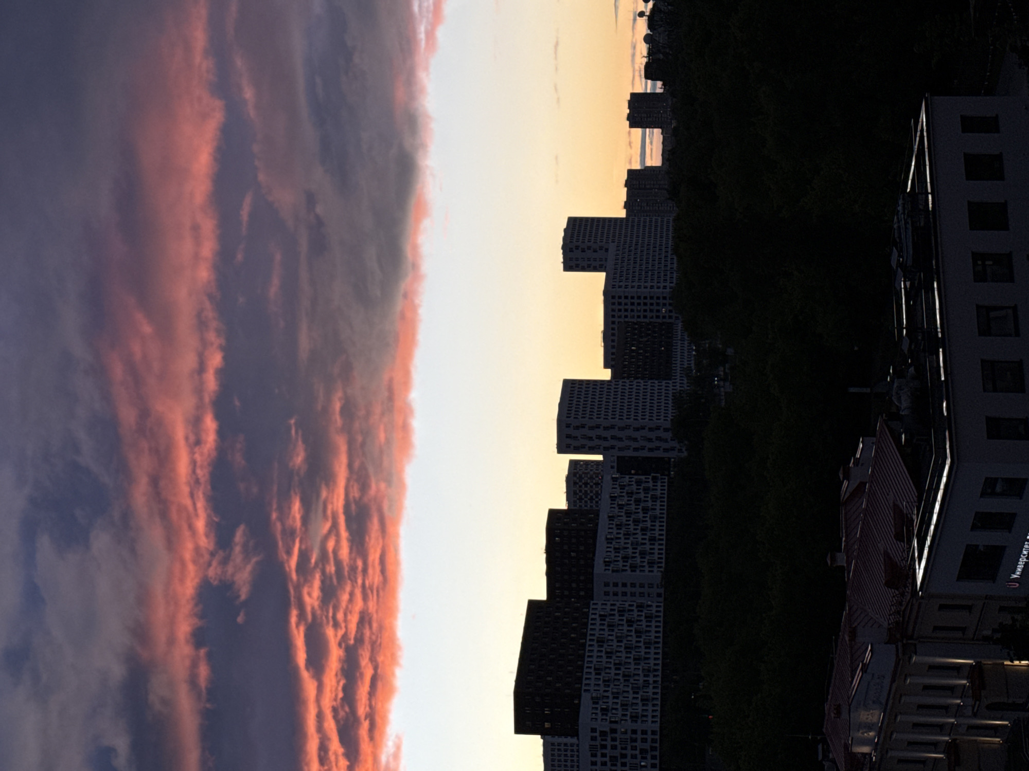 The image shows an urban skyline at sunset in Moscow, Russia. On the left is a dramatic sky filled with pink and purple clouds, suggesting either sunrise or sunset. In the middle ground, a series of modern, high-rise residential buildings punctuate the horizon. To the right, a dark, dense line of trees indicates a park or green space. At the bottom right corner, a building with illuminated windows is visible, with what appears to be Russian text on the side (possibly "Университет" or "University"). The buildings display a regular pattern of windows, indicating modern construction. The dark silhouette of the trees and buildings contrasts sharply with the colorful sky.